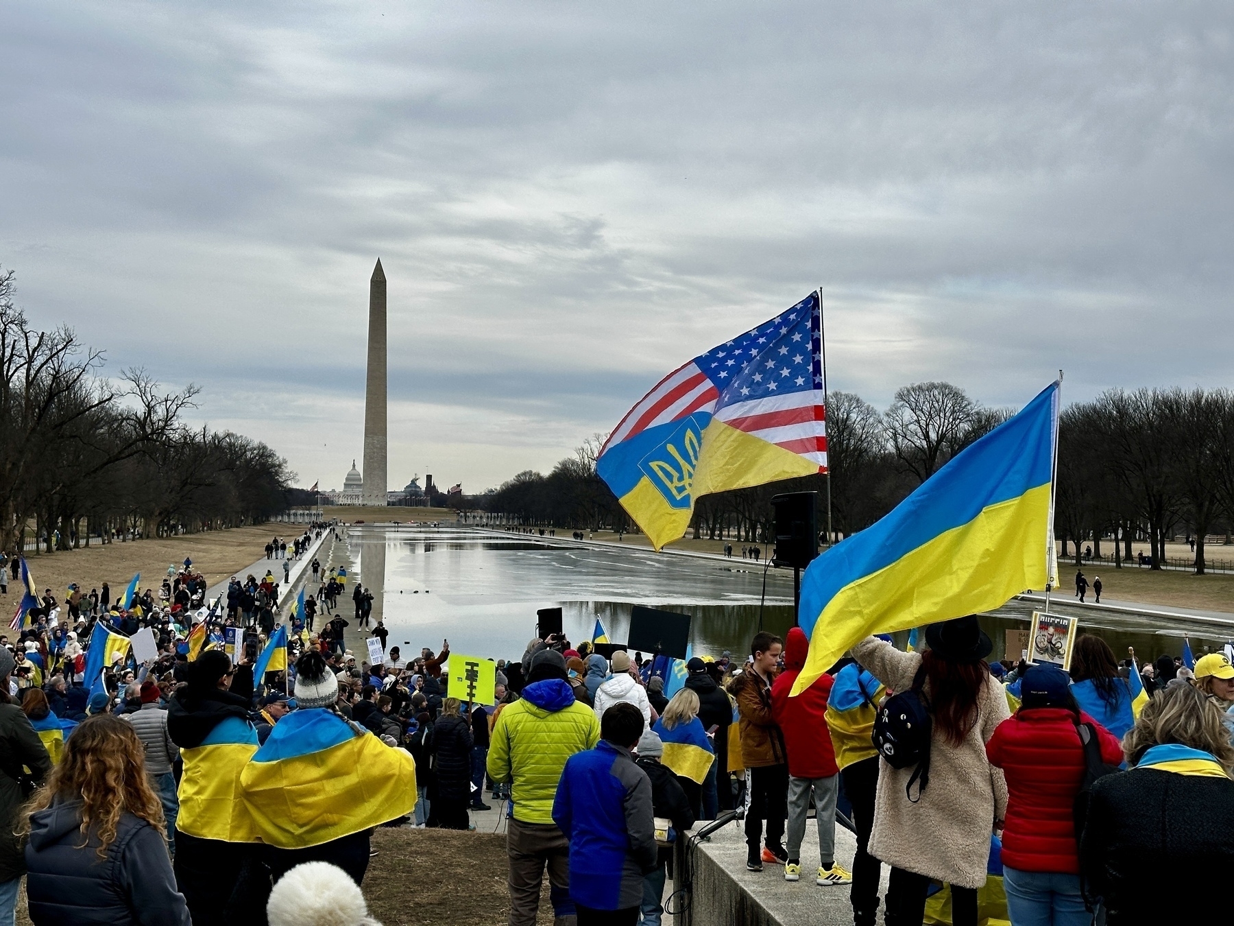 A crowd of people, many holding Ukrainian and American flags, gather near the reflecting pool with the Washington Monument in the background.