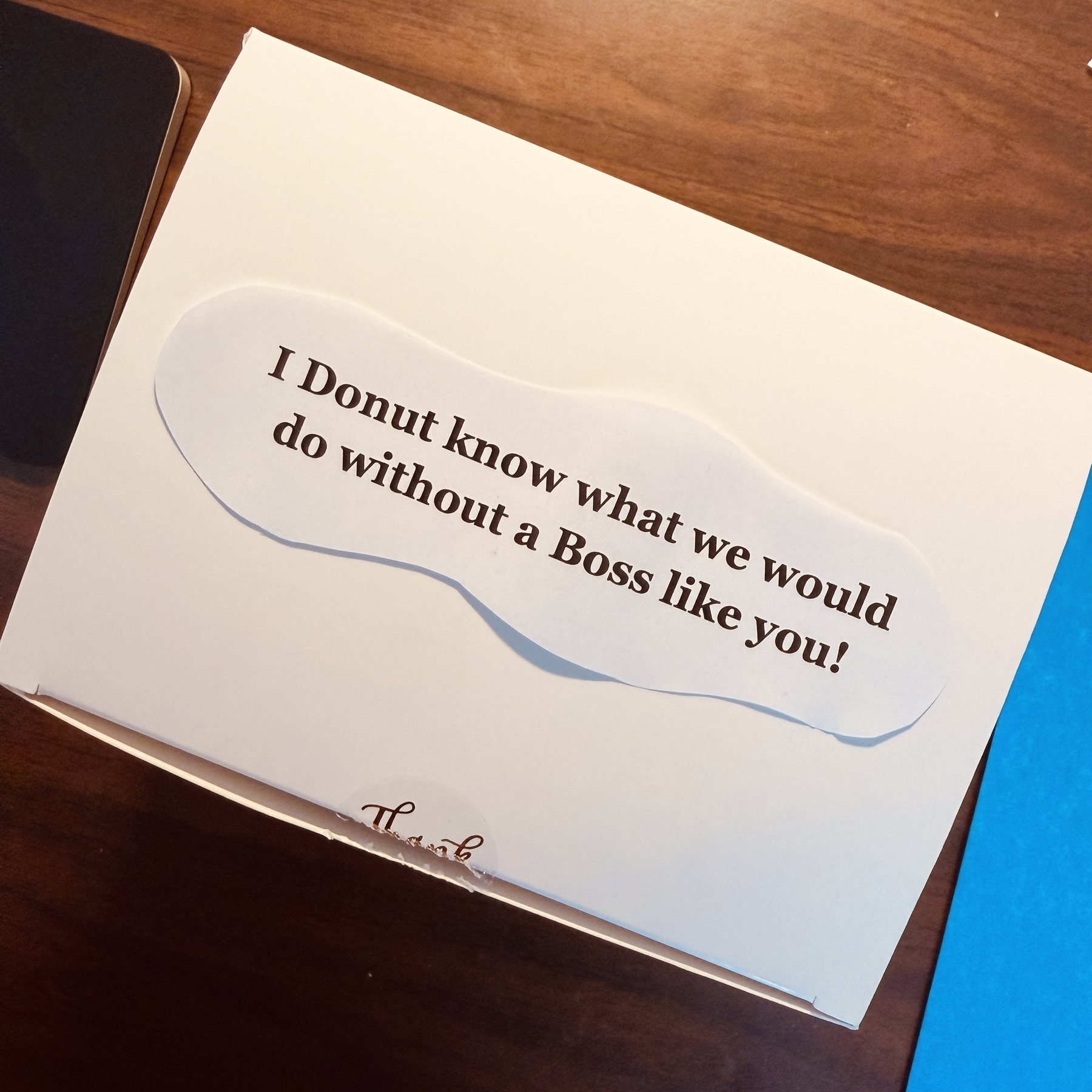 A white box on a brown wooden desk. The label on the box reads, “I Donut know what we would do without a Boss like you!” To the right of the box, part of a blue file folder is visible. To the left of the box, an Apple trackpad is visible.