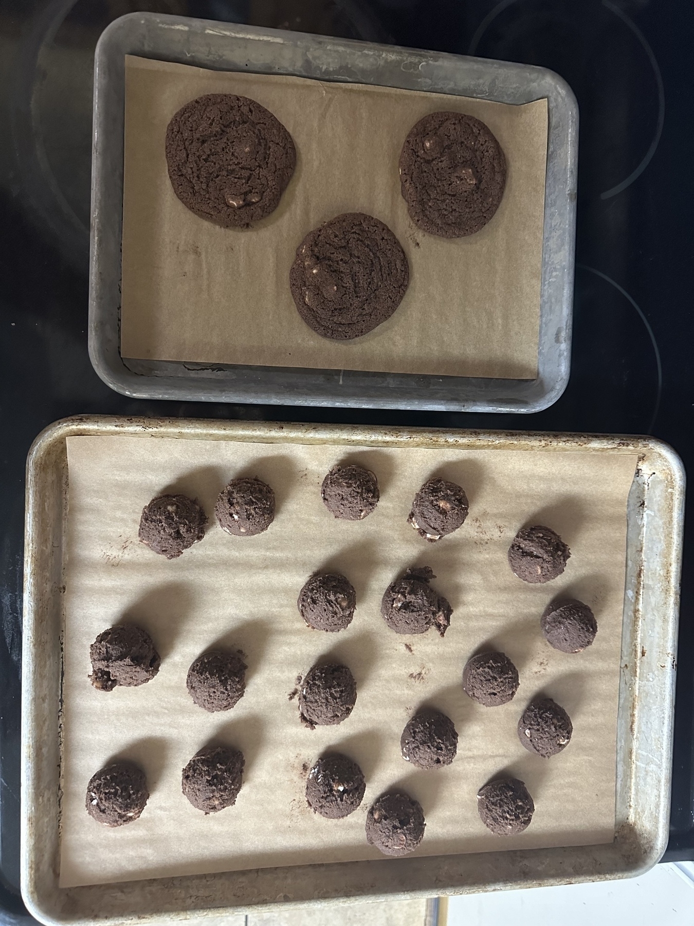 One small tray of baked chocolate cookies next to a larger tray of uncooked dough balls. 