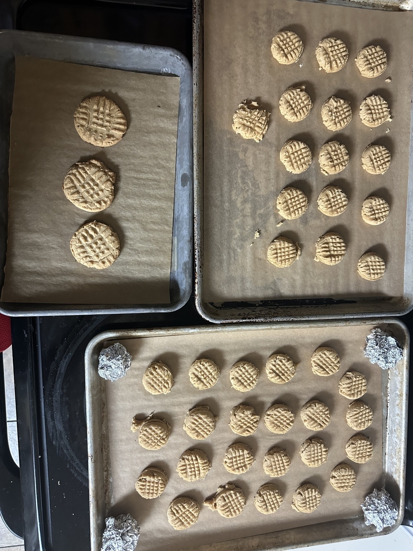Three baked peanut butter cookies on one tray with two other larger trays of unbaked cookies - plus aluminum foil balls that allow the trays to stack in the freezer 