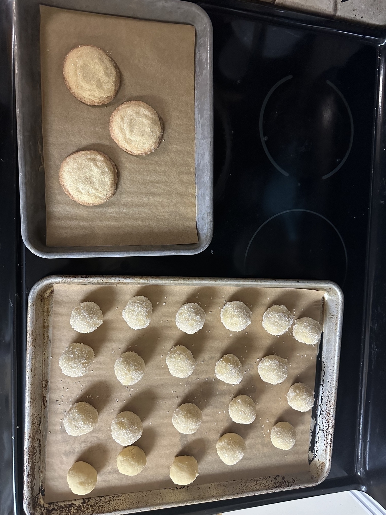 One small tray of cooked sugar cookies next to a bigger tray of unbaked cookies