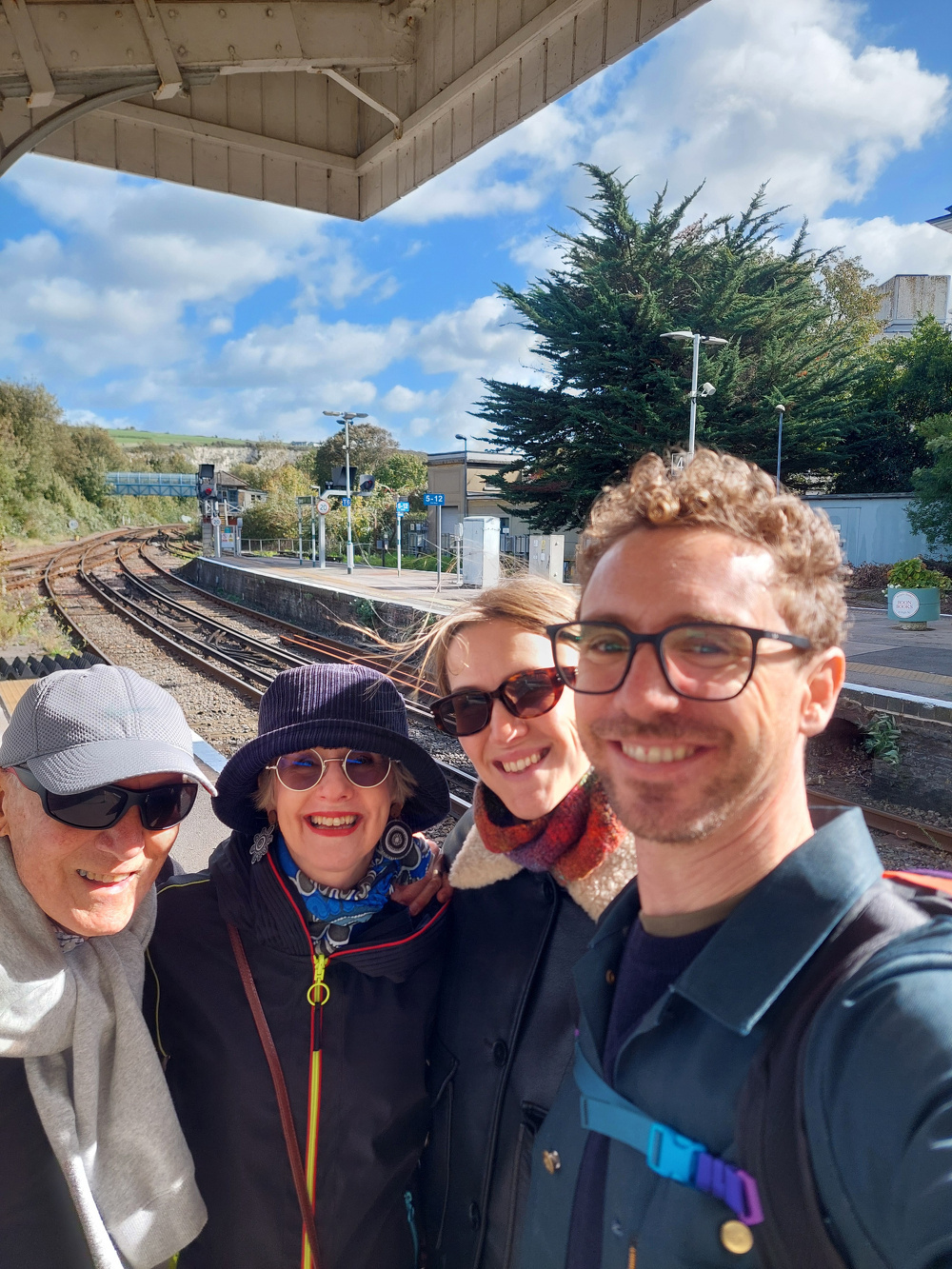 A group of four people is happily posing for a selfie at a sunny train station platform.