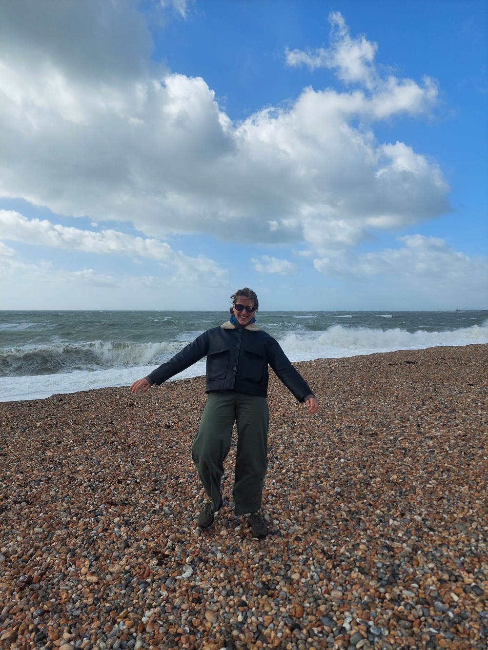 A person is standing on a rocky beach, with waves crashing behind them under a partly cloudy sky.