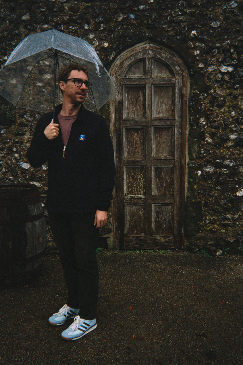 A person stands holding a transparent umbrella in front of a rustic stone wall and wooden door.