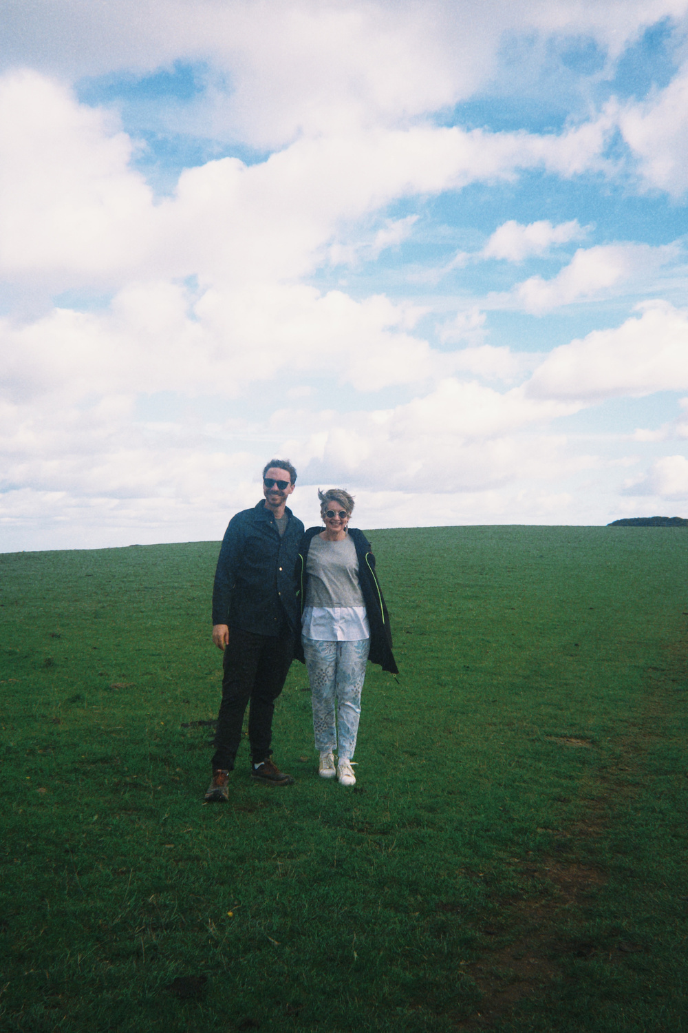 A couple stands together smiling on a vast, open grassy field under a partly cloudy sky.