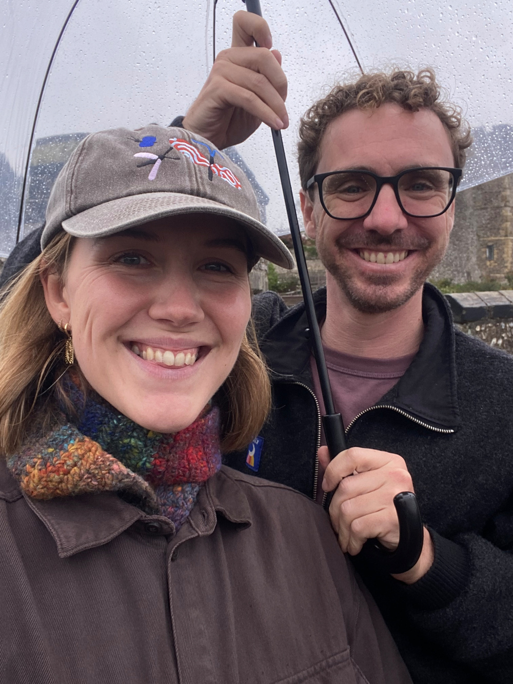 Two people smiling under a transparent umbrella on a rainy day.