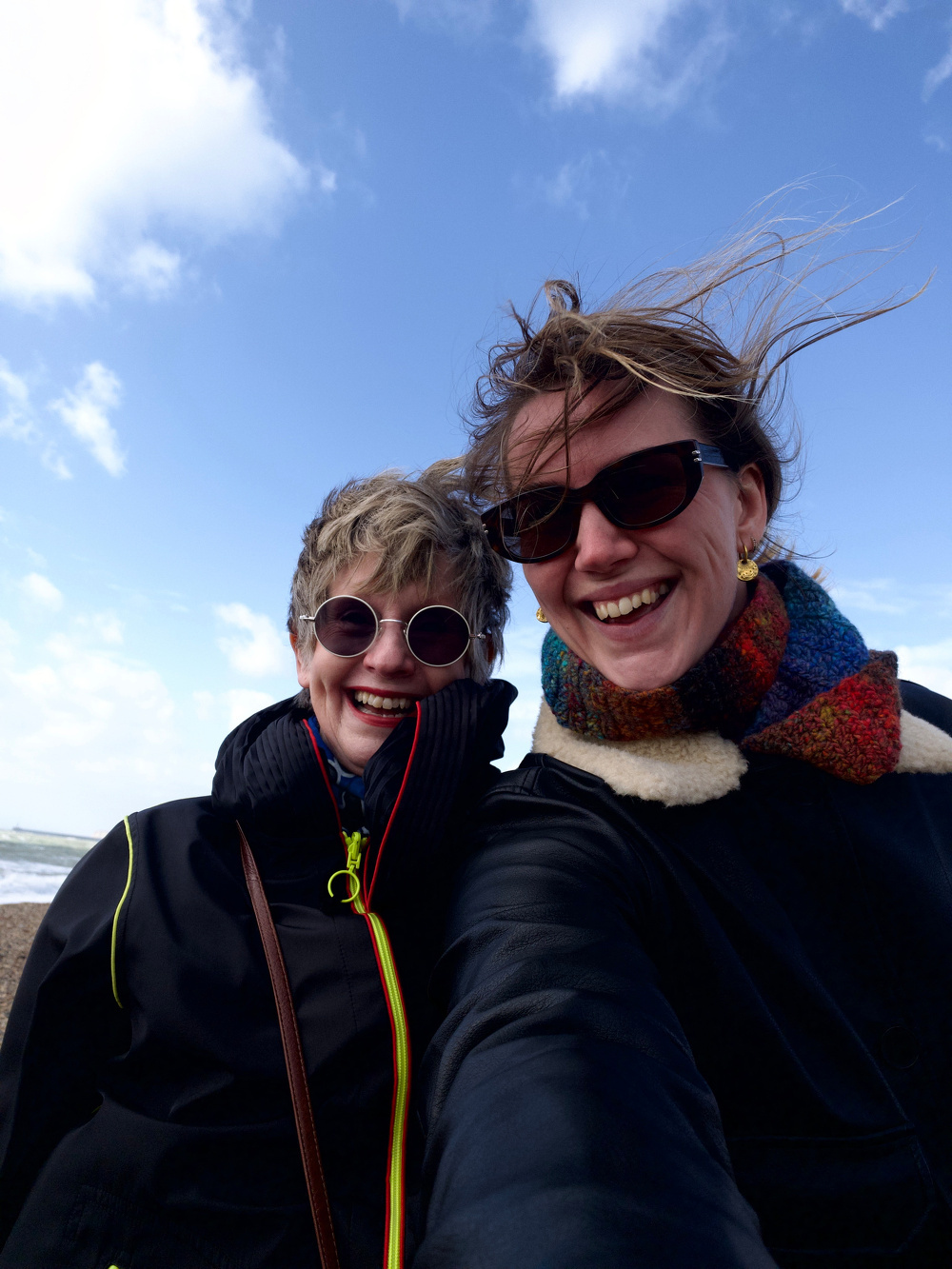 Two people wearing sunglasses and jackets smile for a selfie on a windy beach day.