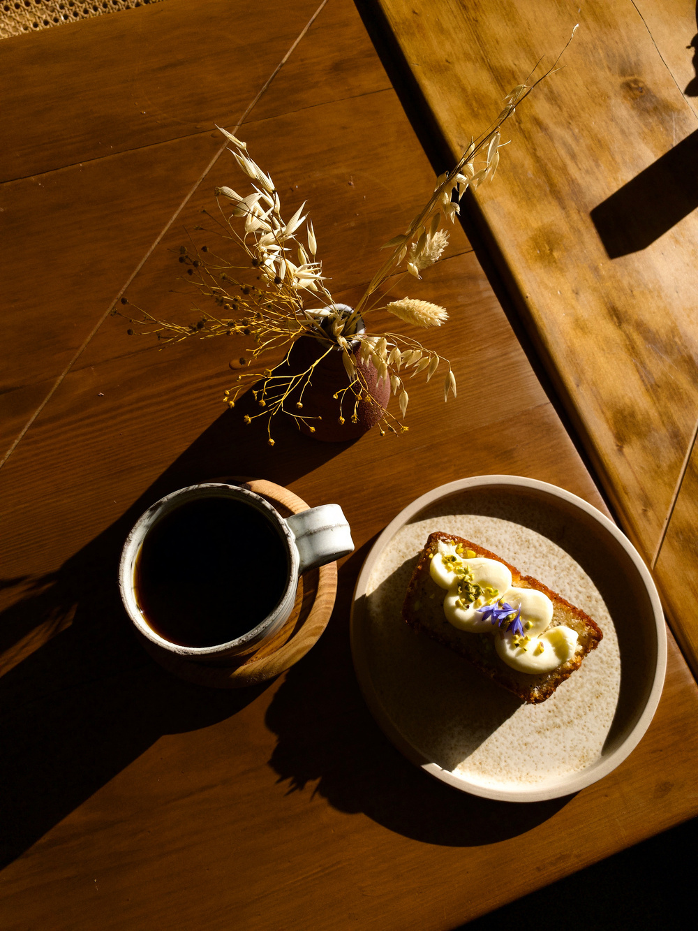 A cup of coffee and a slice of bread topped with sliced plums and garnished with a flower are placed on a wooden table, alongside a vase with dried flowers.