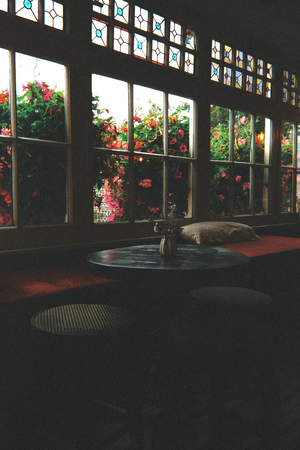 A cozy interior features a round table and stools by a row of large windows adorned with vibrant hanging flowers outside.