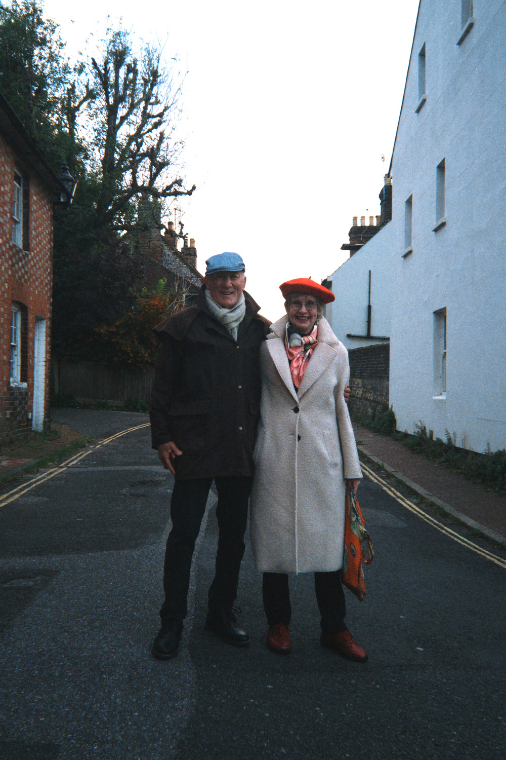 Two people are smiling and posing together on a quiet street, wearing coats and hats.