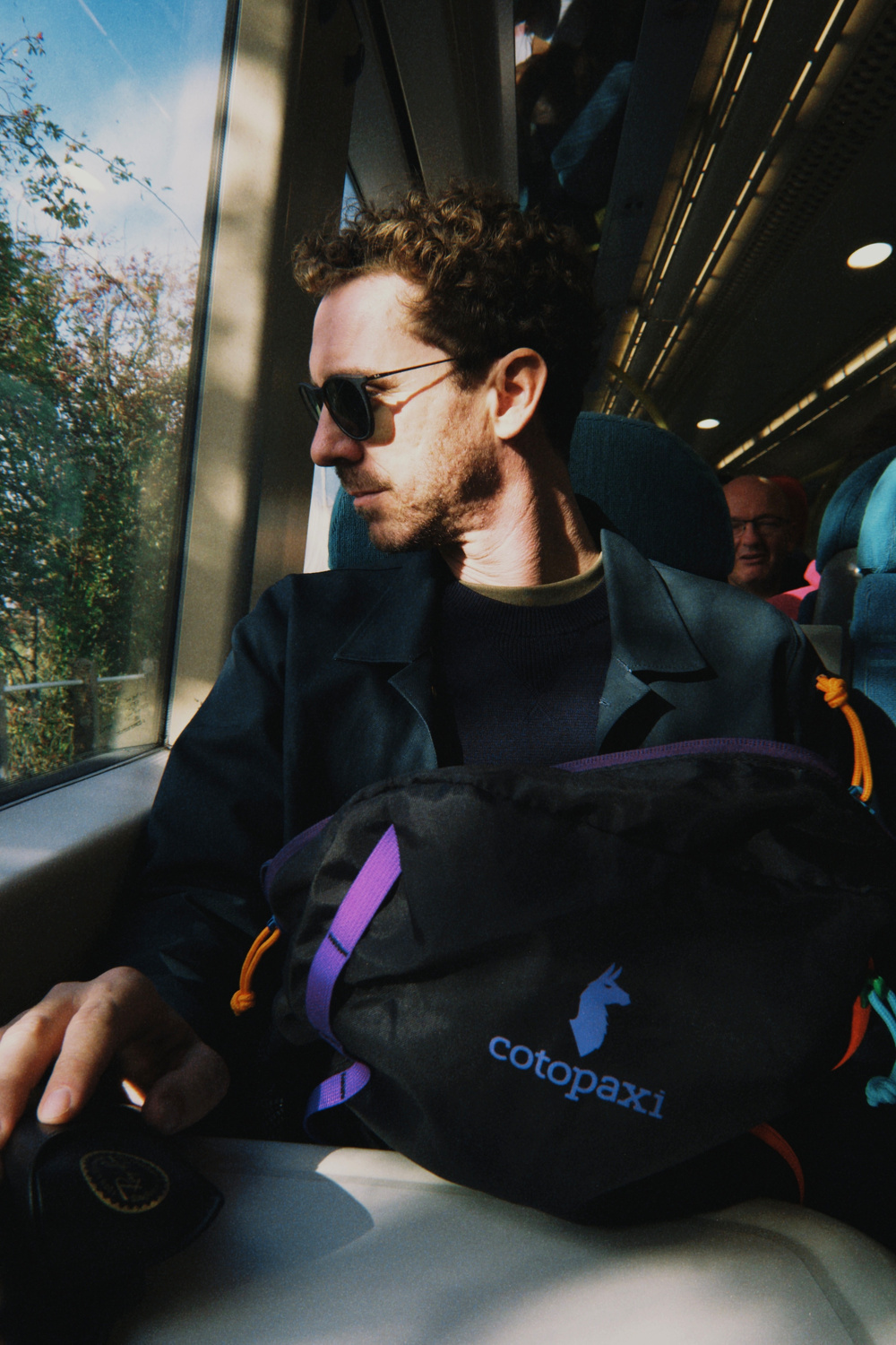 A man wearing sunglasses sits on a train, looking out the window while holding a bag labeled cotopaxi.