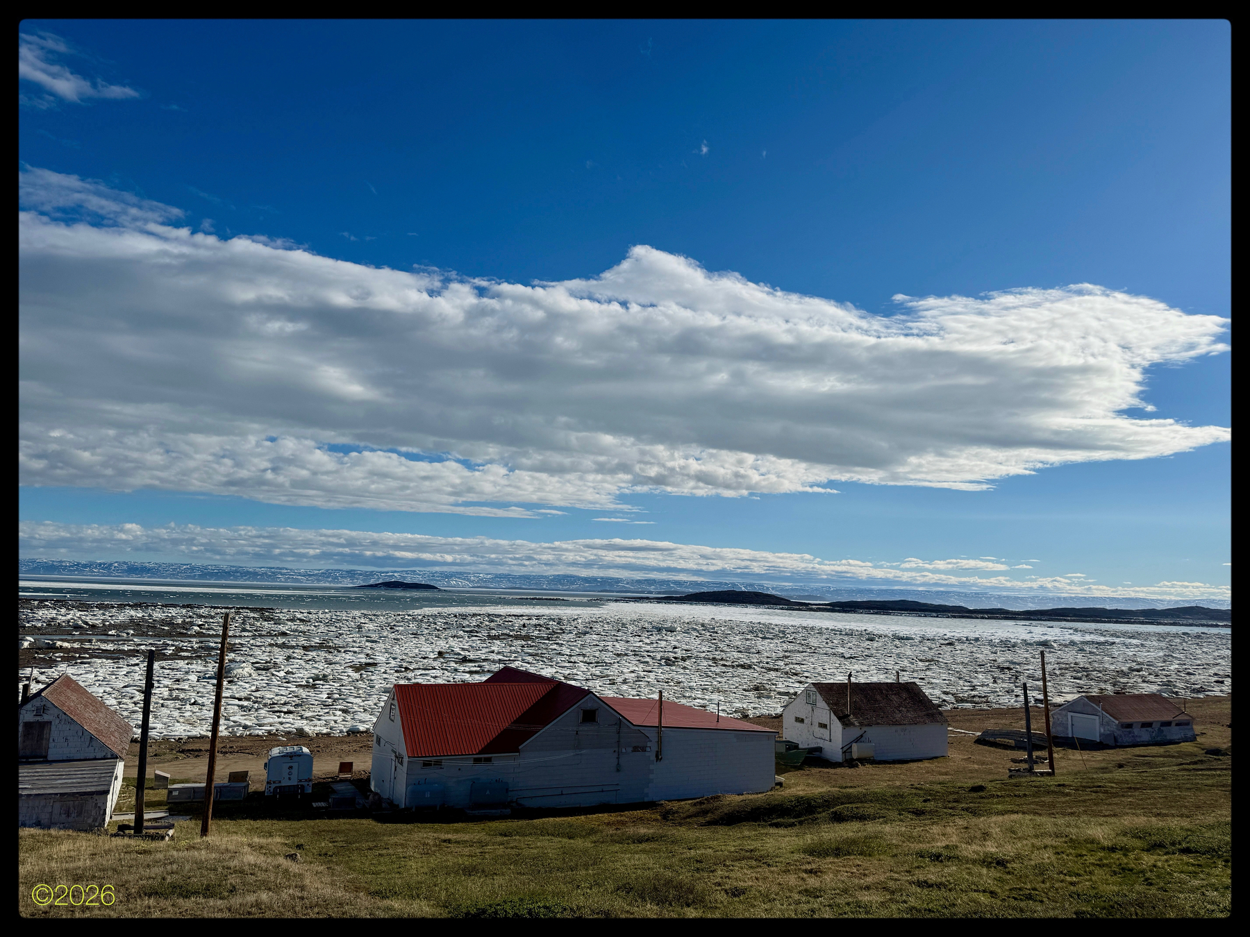 A few small HBC houses with red & white roofs are situated on a sandy shore overlooking a mostly ice-covered body of water under a bright, partially cloudy sky.