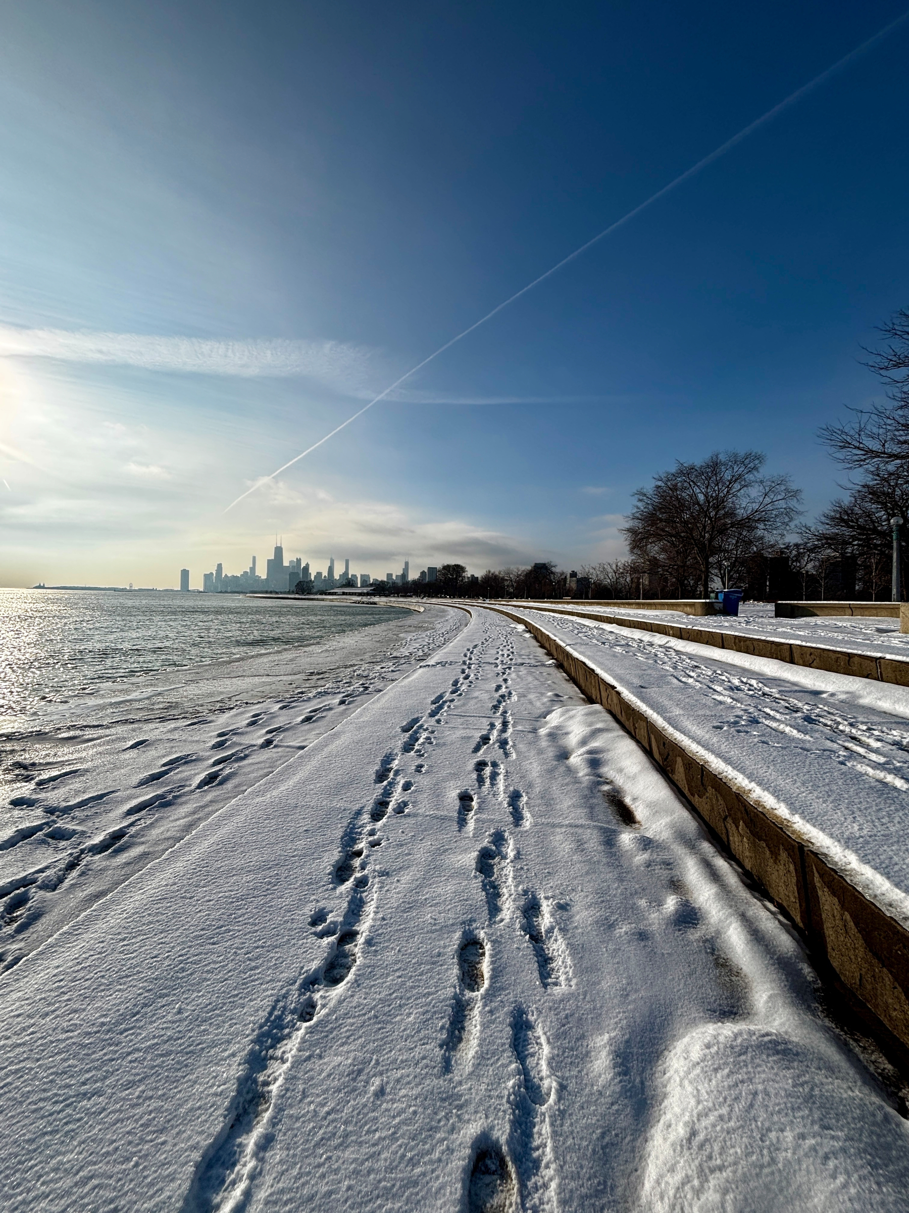 A snowy pathway with footprints stretches alongside Lake Michigan in the morning, leading towards a city skyline under a clear blue sky.