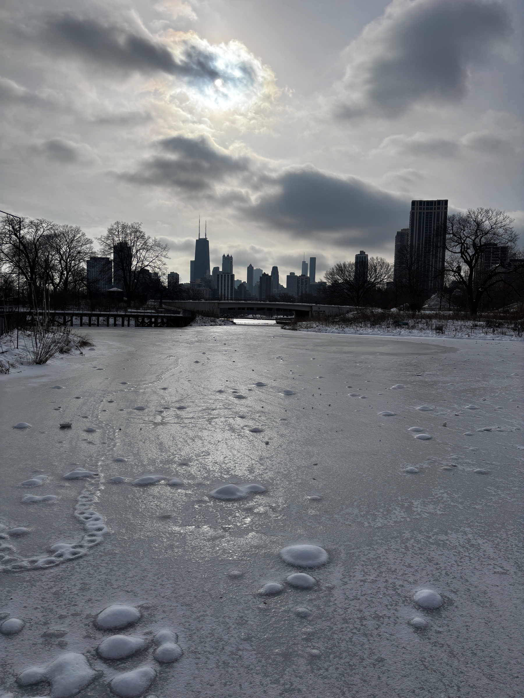 A frozen body of water with scattered bubbles is set against a city skyline under a dramatic, cloudy sky.