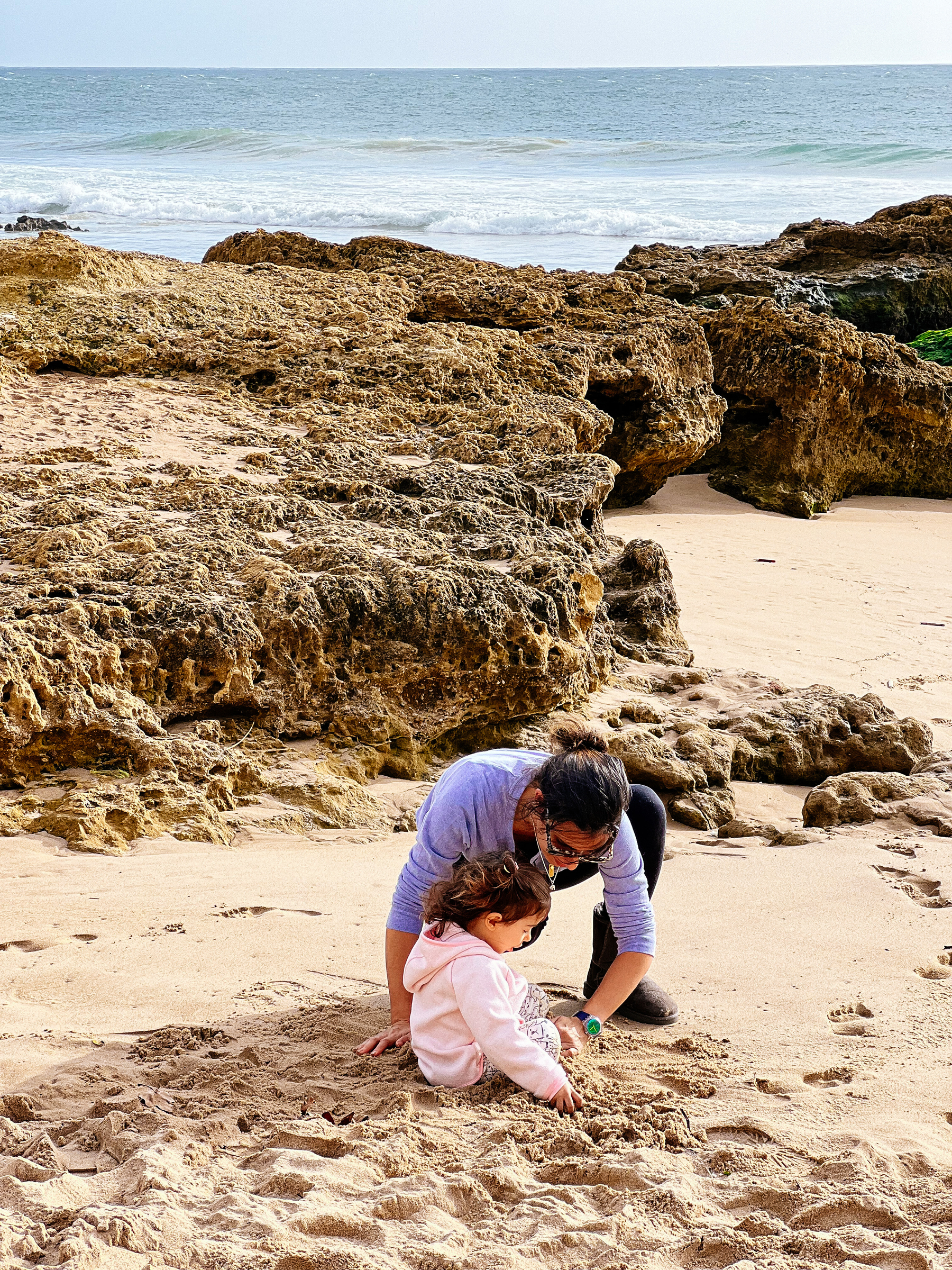 A mom plays with a toddler in the sand, sea behind them. 