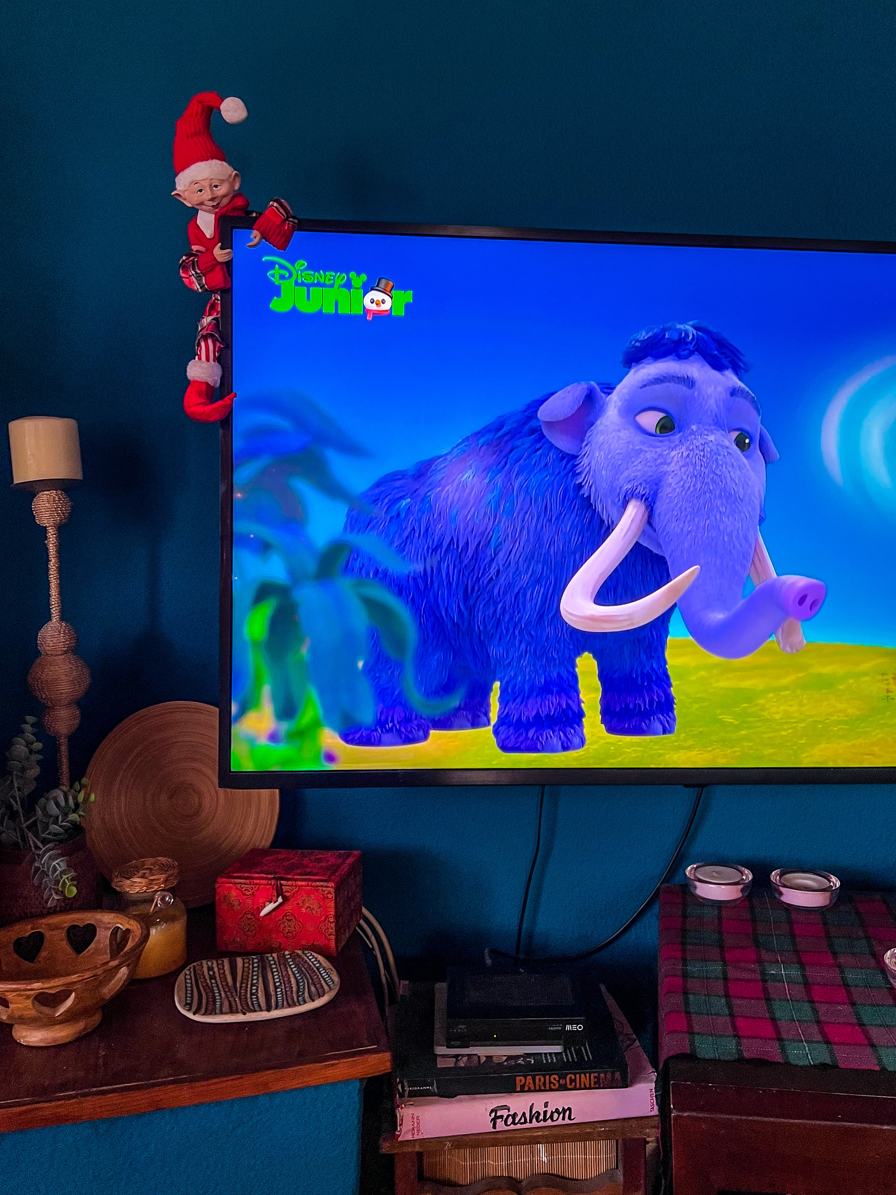 A tv on Disney Junior channel, with a blue mammoth playing. A tiny elf is hanging from the side of the tv. 
