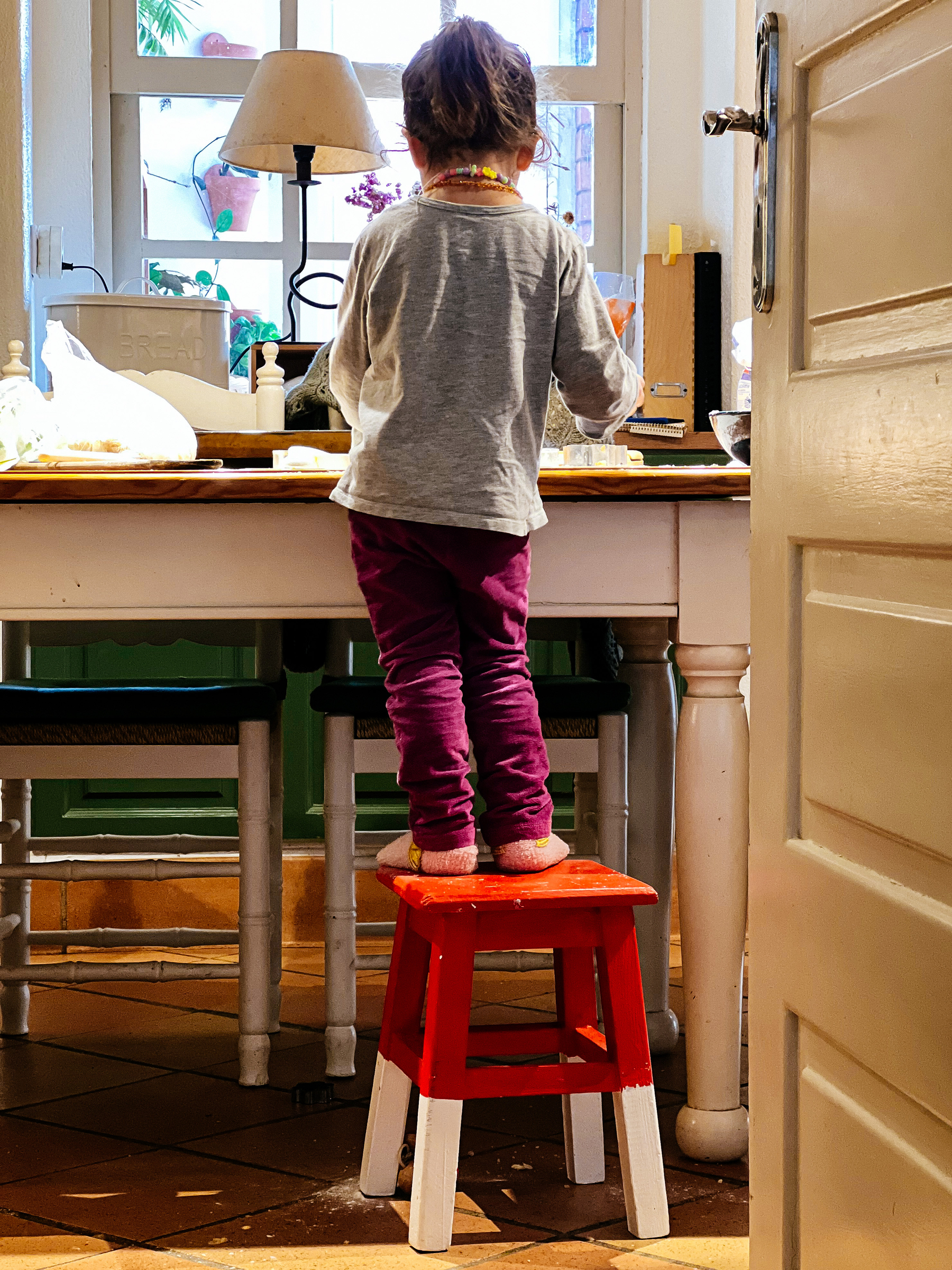 Toddler baking, photo from her back, standing on a stool. 