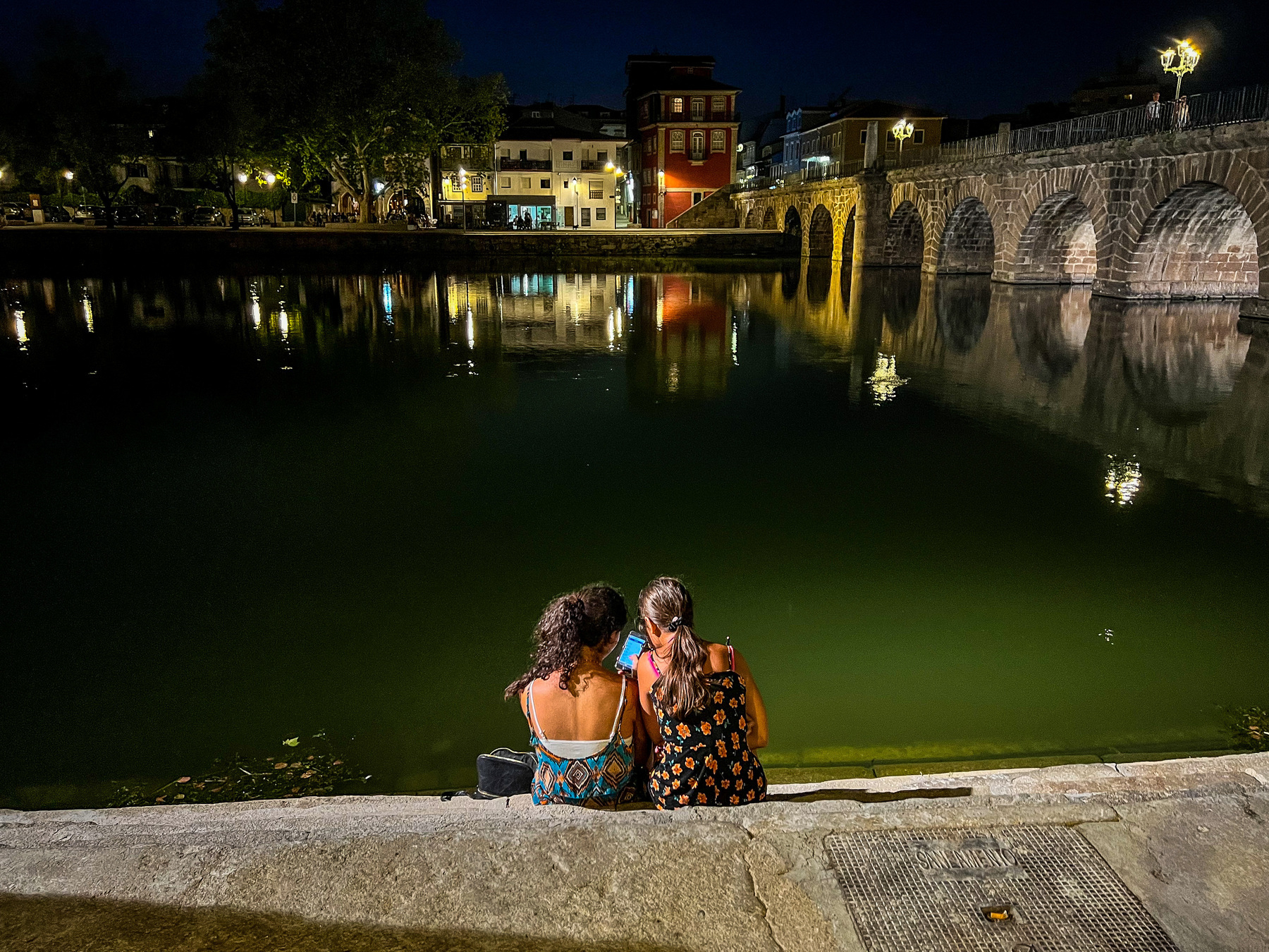 Two girls watch a phone, with a Roman bridge in the background 