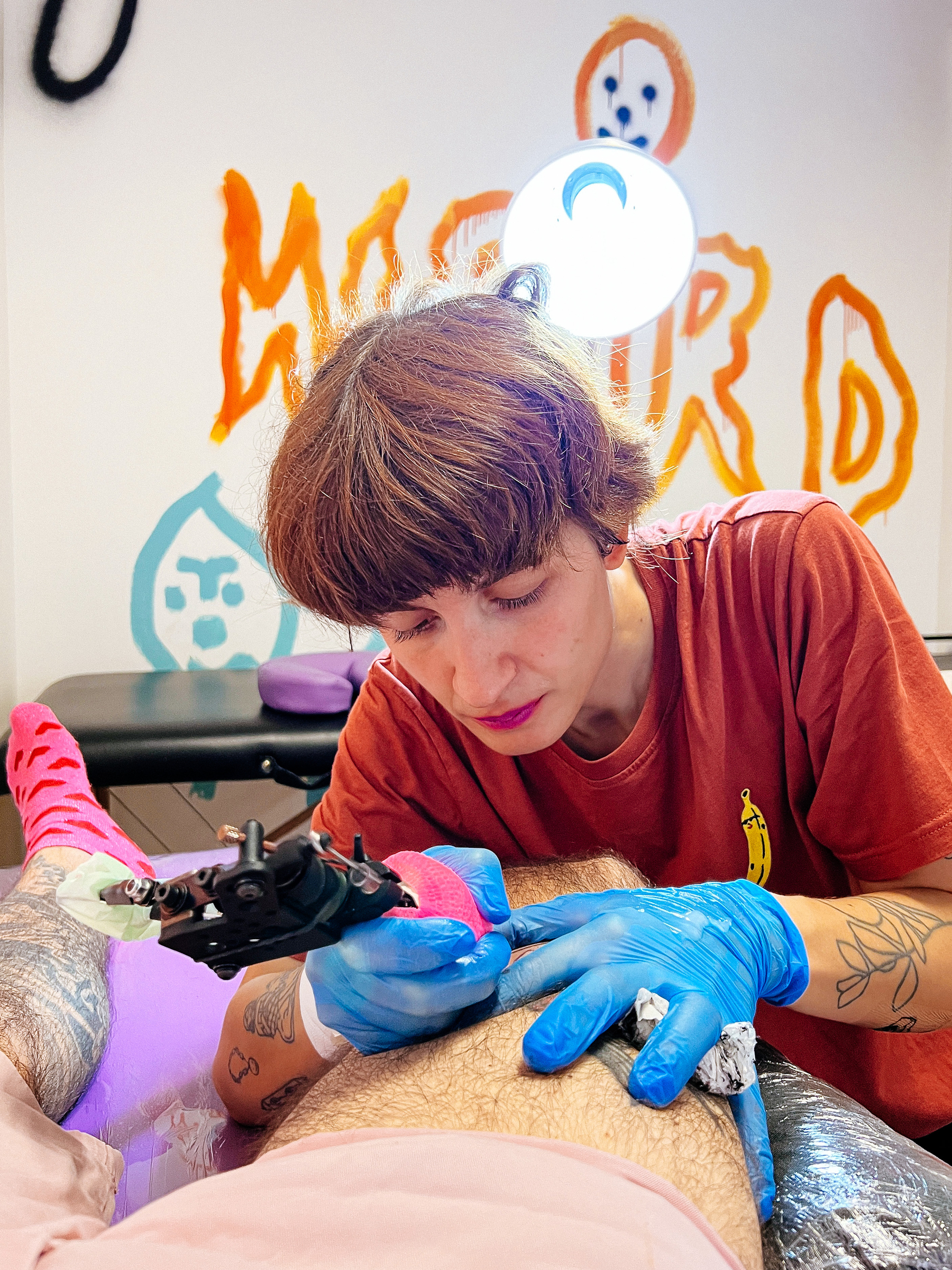 A girl tattoo artist works on a leg. “Weird” written on the wall behind her. 