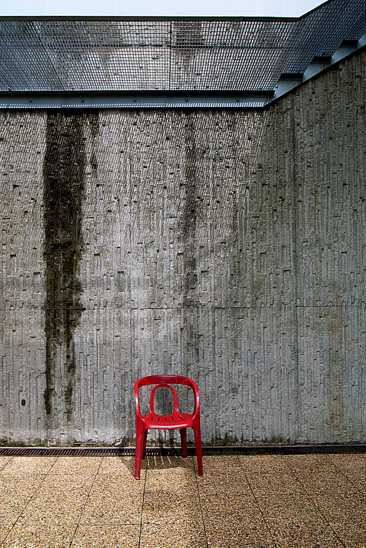 A lonely red plastic chair sits against a very tall grey wall. 