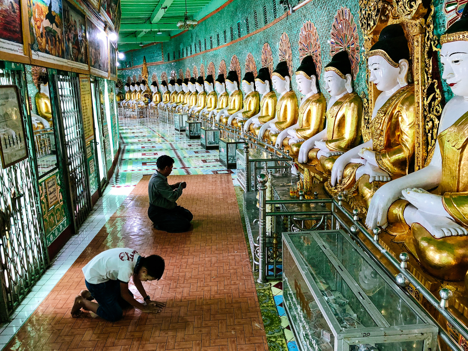 People pray inside a temple in Mandalay. The temple is mostly green, with a row of Buddha statues on the right side. 