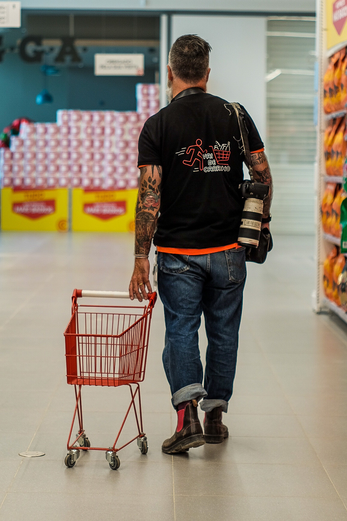 A man walks down a supermarket isle with a tiny cart in tow. 