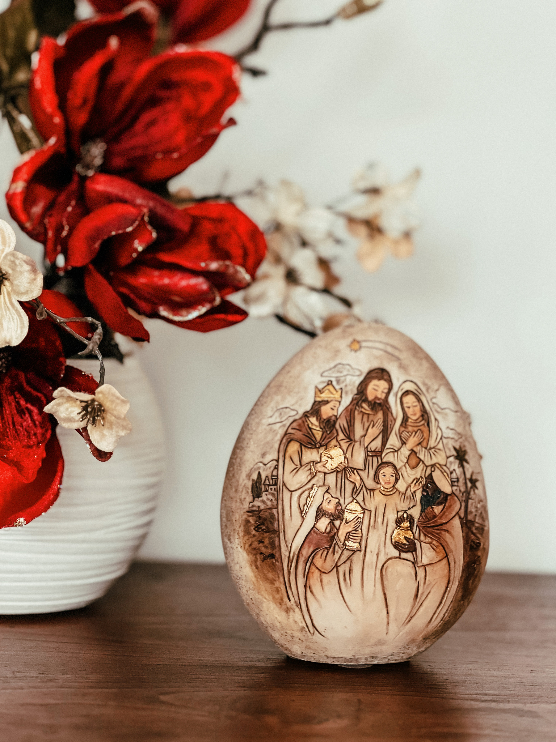 A stone “egg” with a nativity scene painted on it. On the left side a vase with red flowers. 