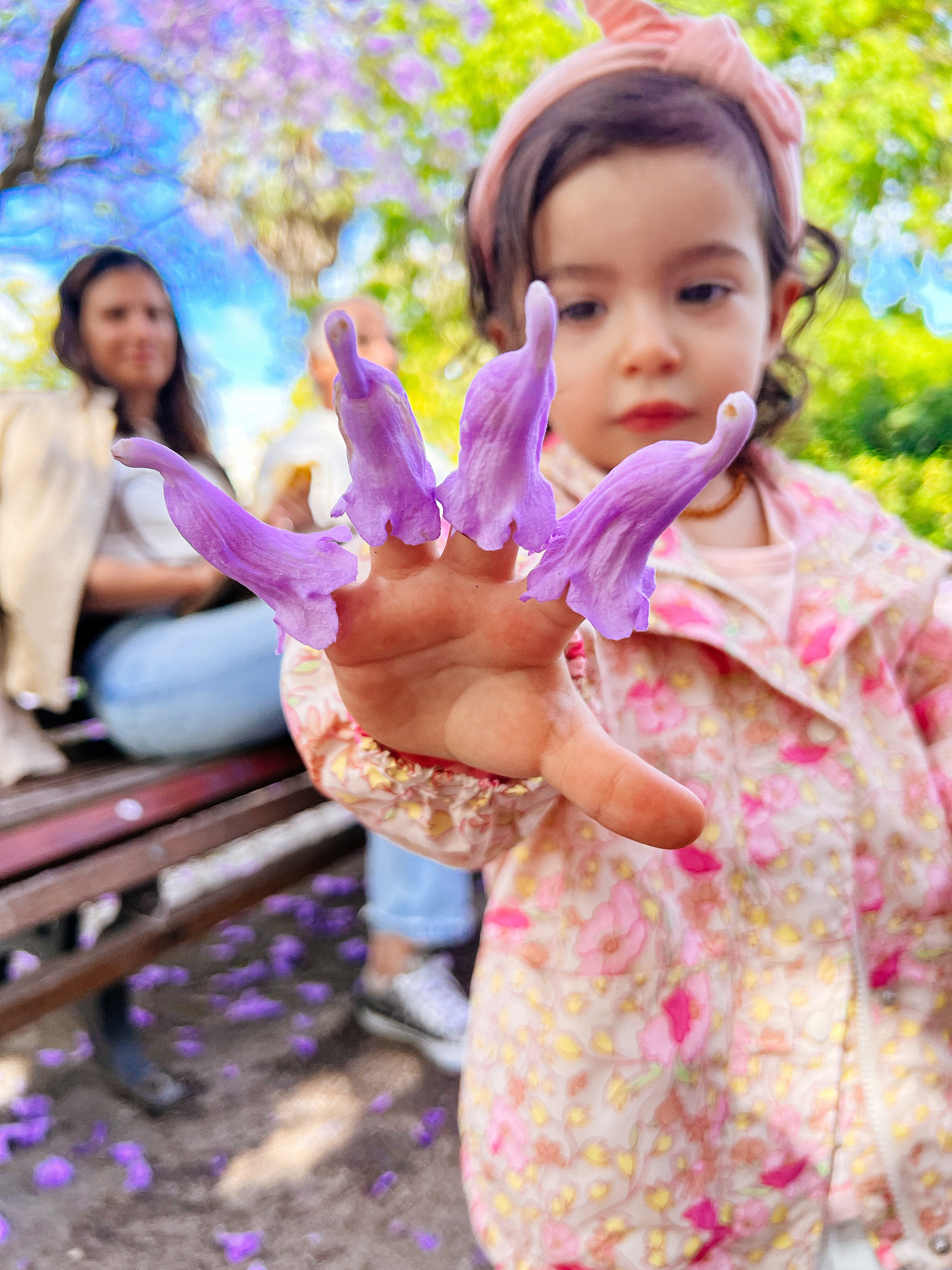 A toddler showing her hand, with a jacaranda flower on each finger. In the back her mom smiles, out of focus. 