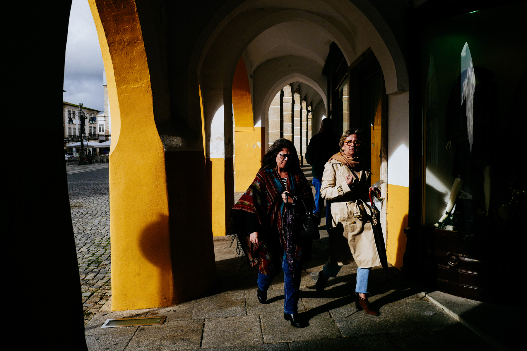 Two women walk towards us, under yellow arches. 