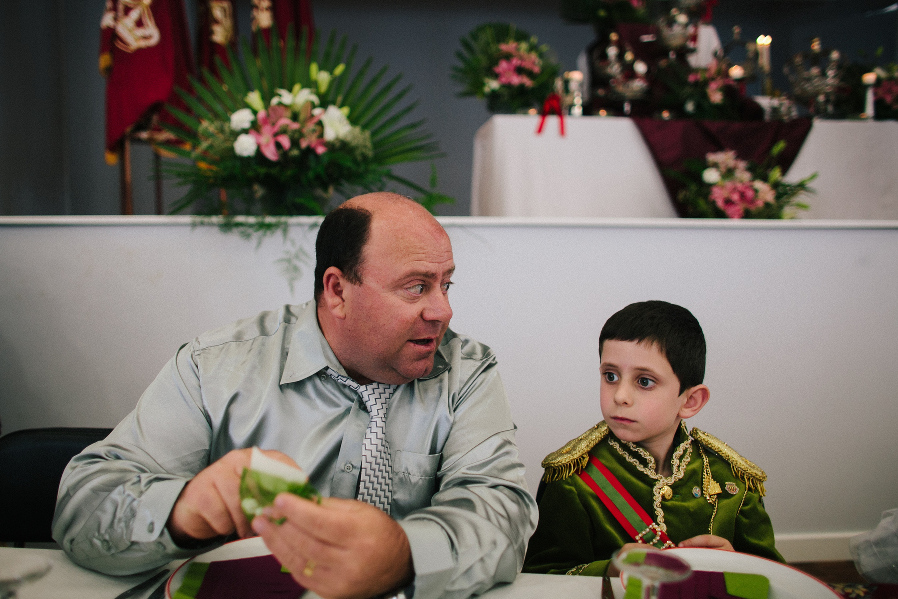 A boy sits next to a man at a lunch table. The boy is wearing a sort of pretend uniform, in green, and looks very serious. 