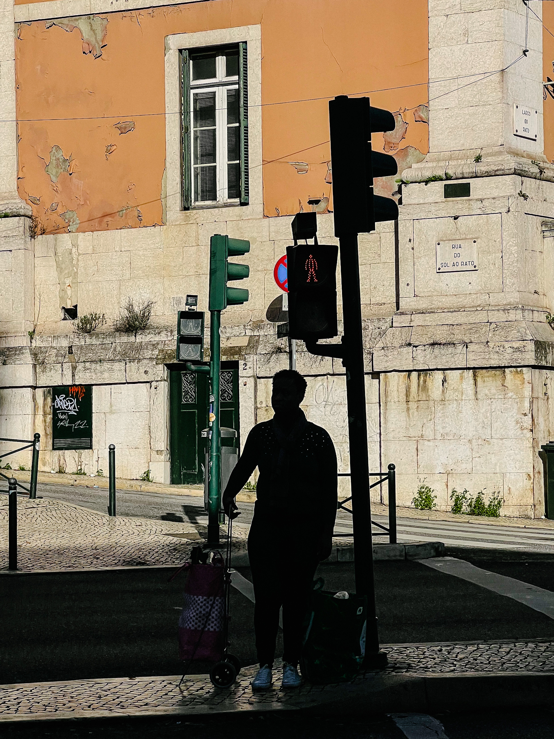 A woman waits for the green light to cross the street. We can see an old building in the back and traffic lights, one with a red light. 