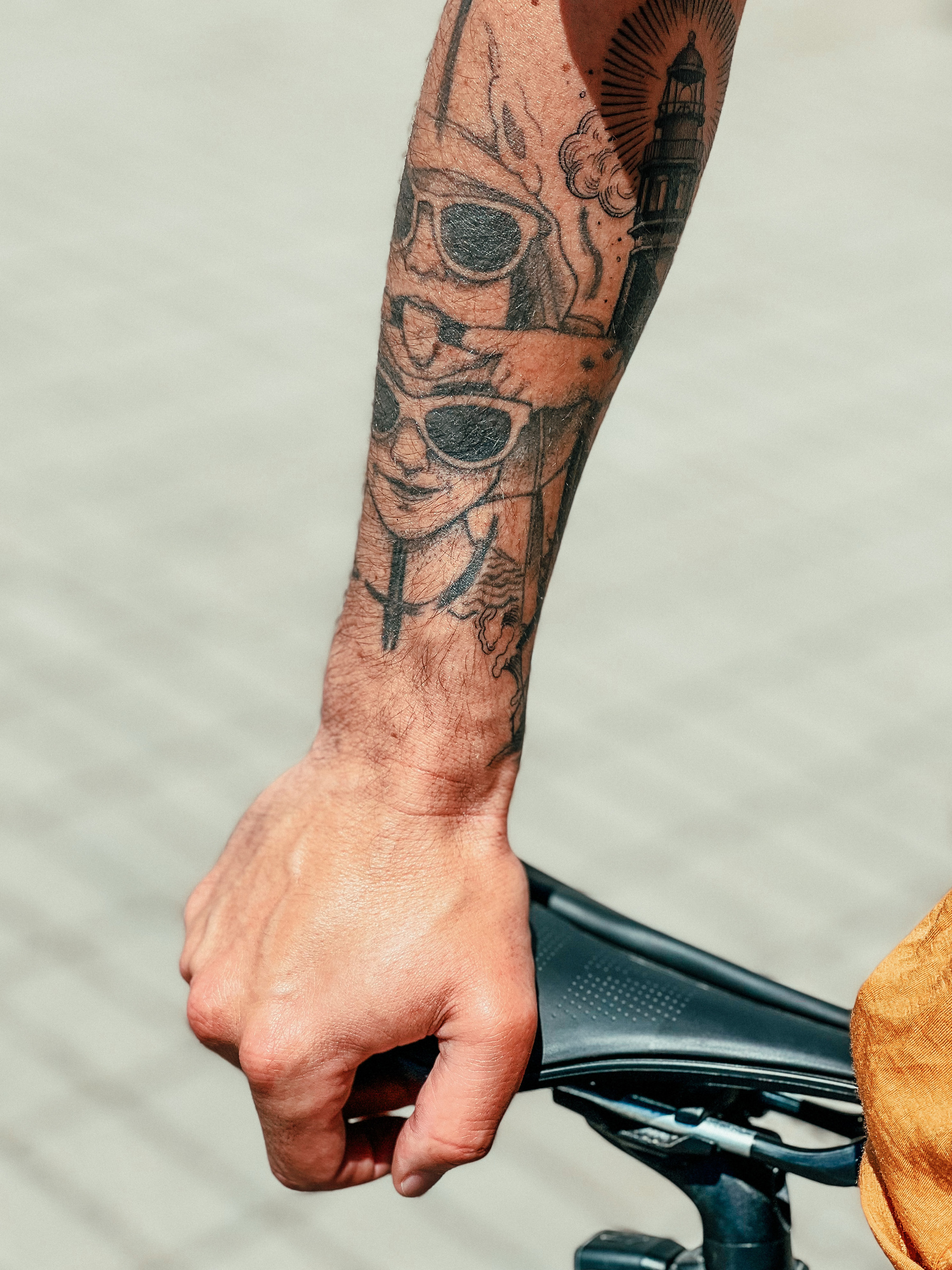 A hand rests on the seat of a bicycle. On the arm you can see a tattoo depicting two kids, and a lighthouse. 