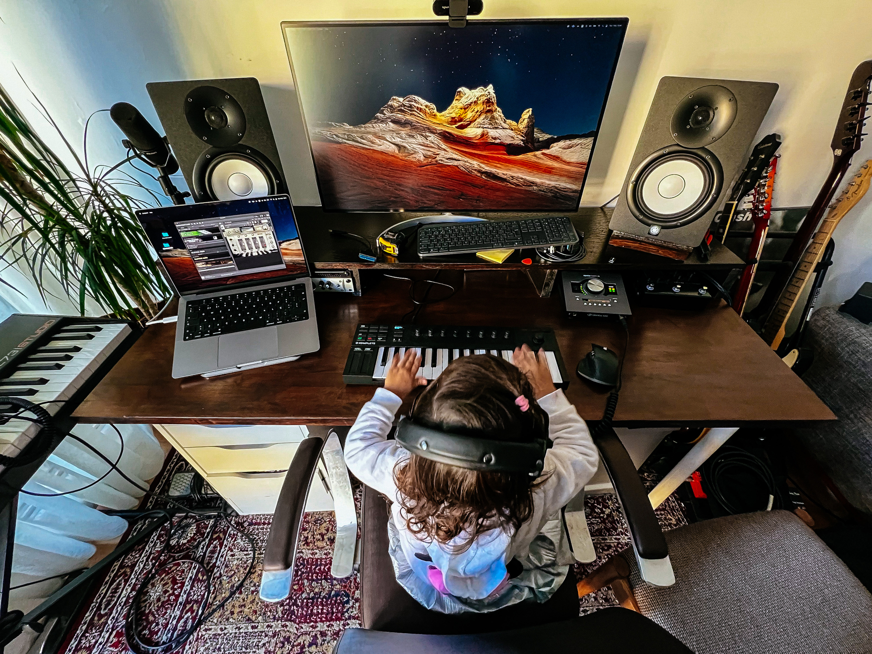 A kid sitting down in front of a piano keyboard, and a table full of computers, speakers, guitars…