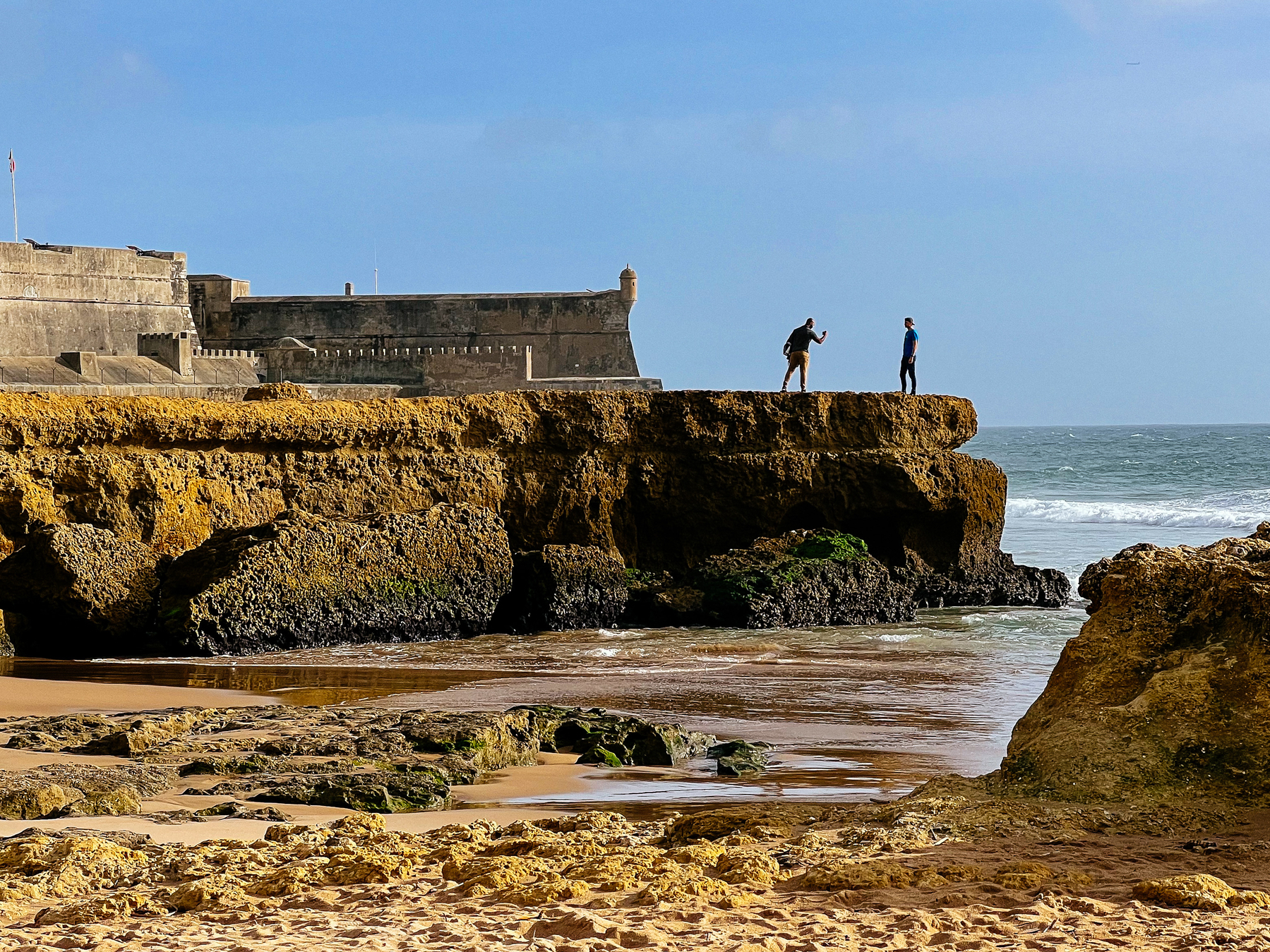 Men takes photos by the sea, with a fort in the background 