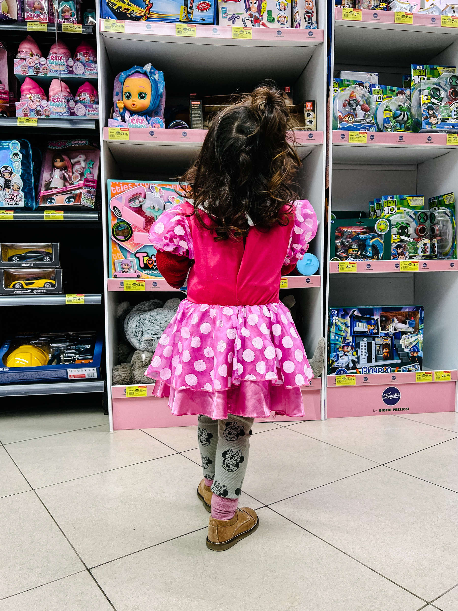 A toddler seen from the back, dressed as Minnie Mouse. She’s looking at a rack full of toys. 