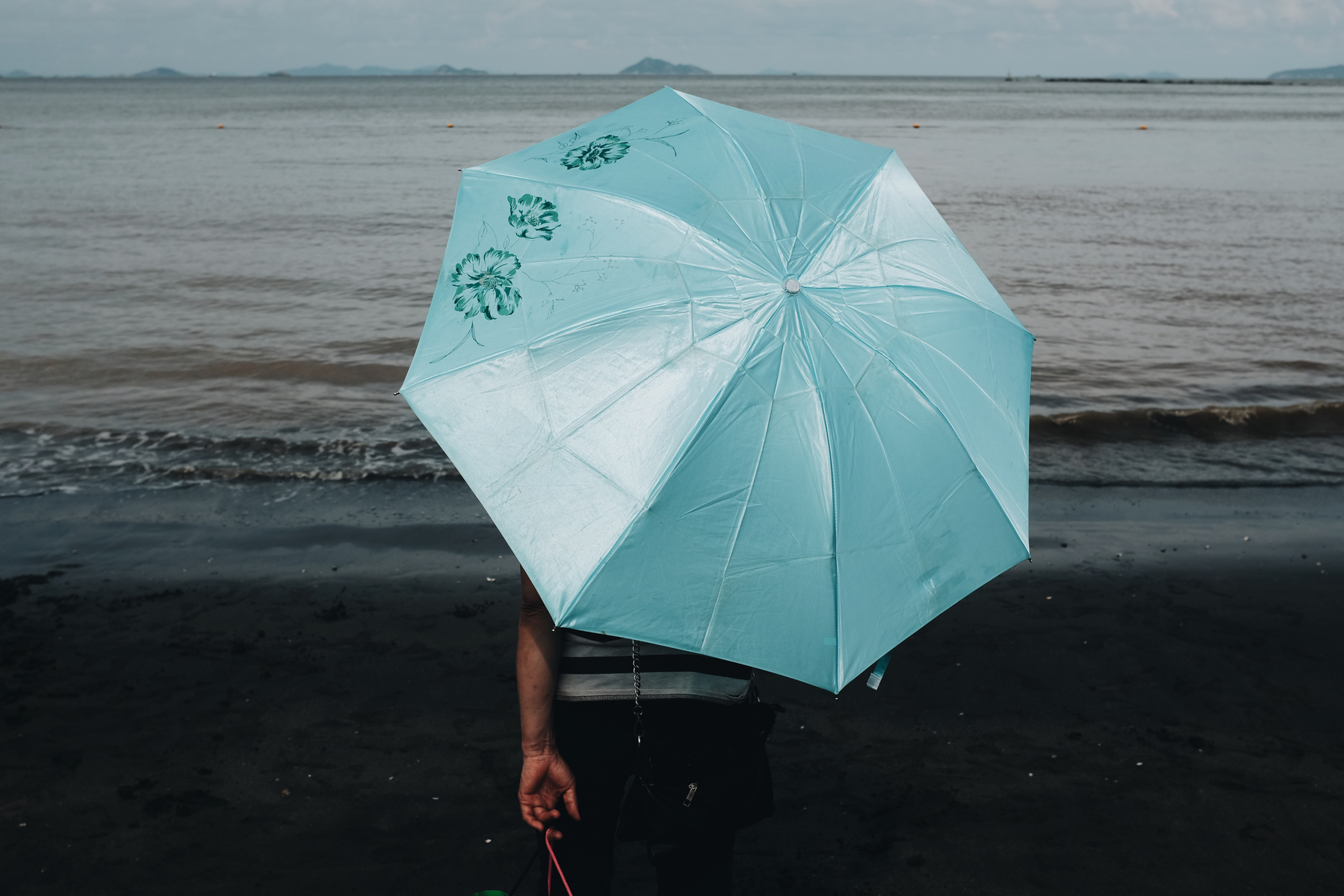 A person stands close to the water with a blue umbrella. The sand on the beach is black. 
