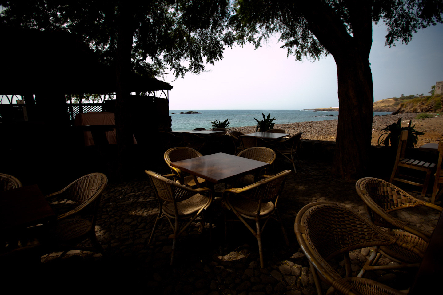 Chairs and tables sit under trees, next to a beach. 