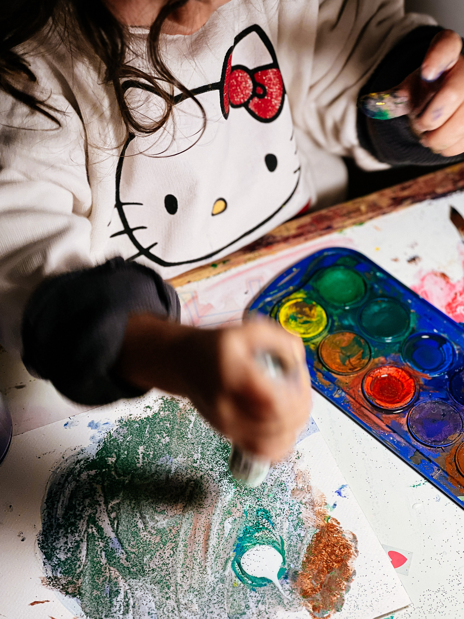 A toddler plays with paint and glue wearing a Hello Kitty sweatshirt. 