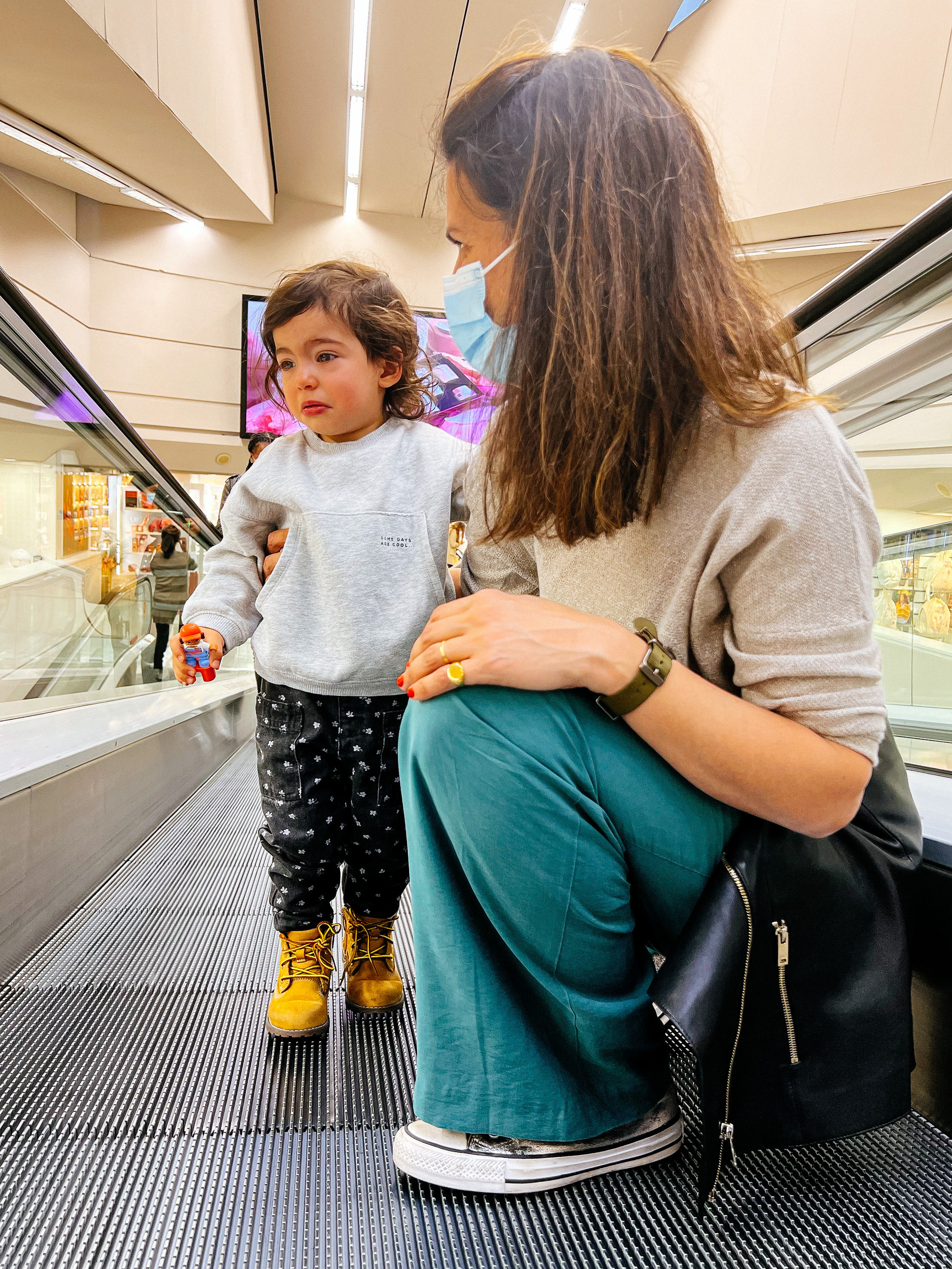 A toddler and her mom ride a moving walkway. The mom is on her knee, the little one seems a little distressed by the experience. 
