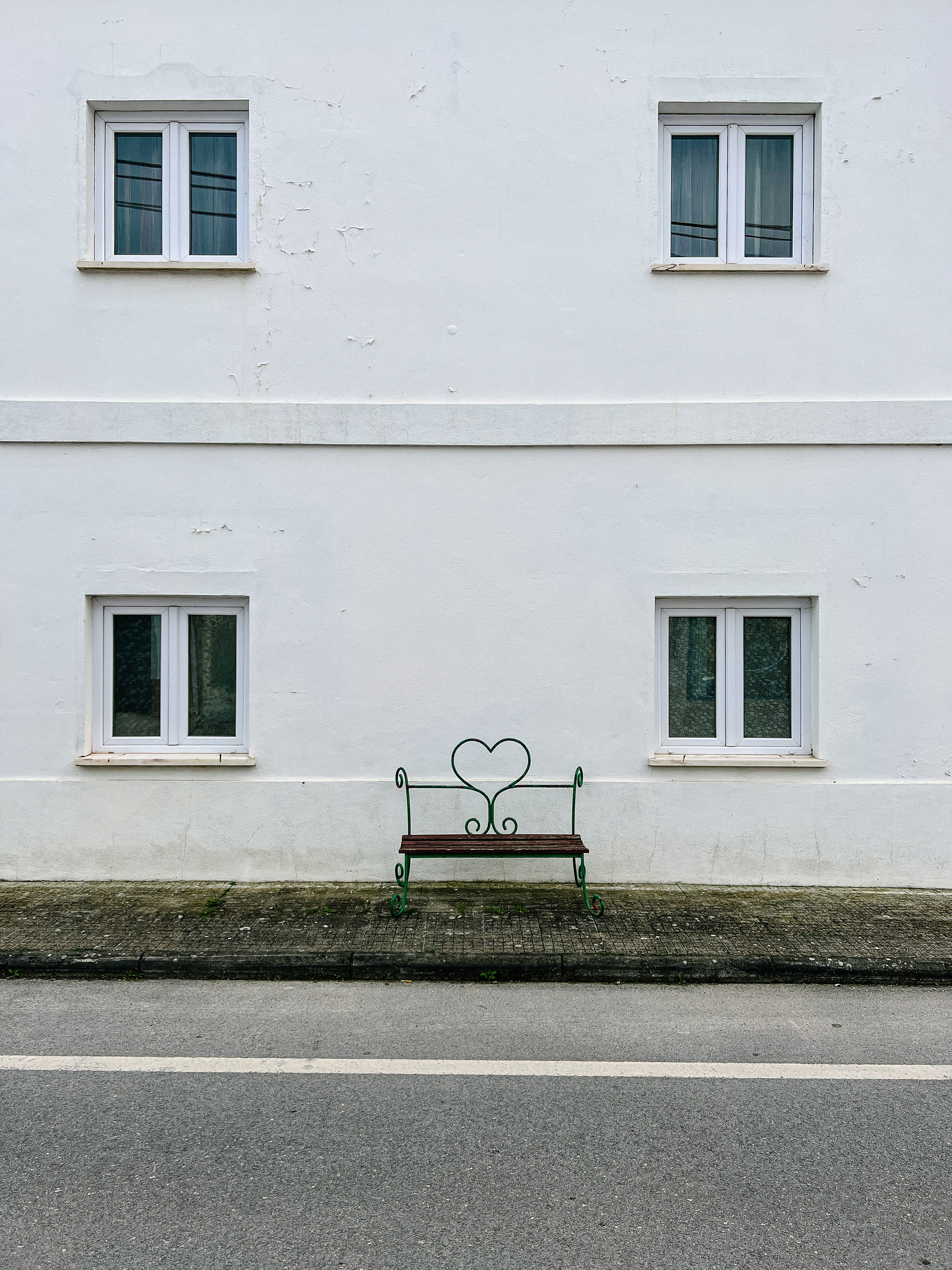A bench with a heart sits against a white wall with four windows.