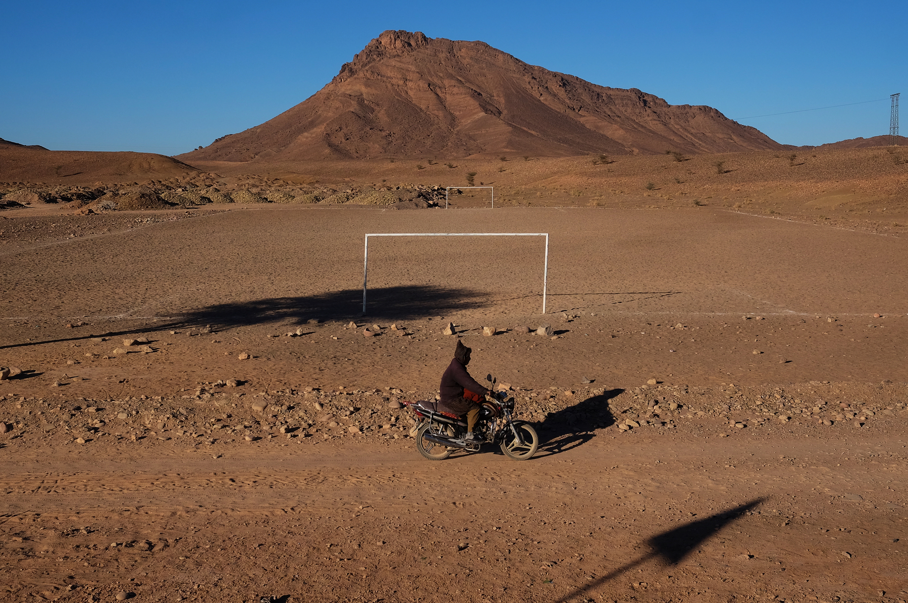 A man rides a motorcycle in the desert. Behind him two football goals. 