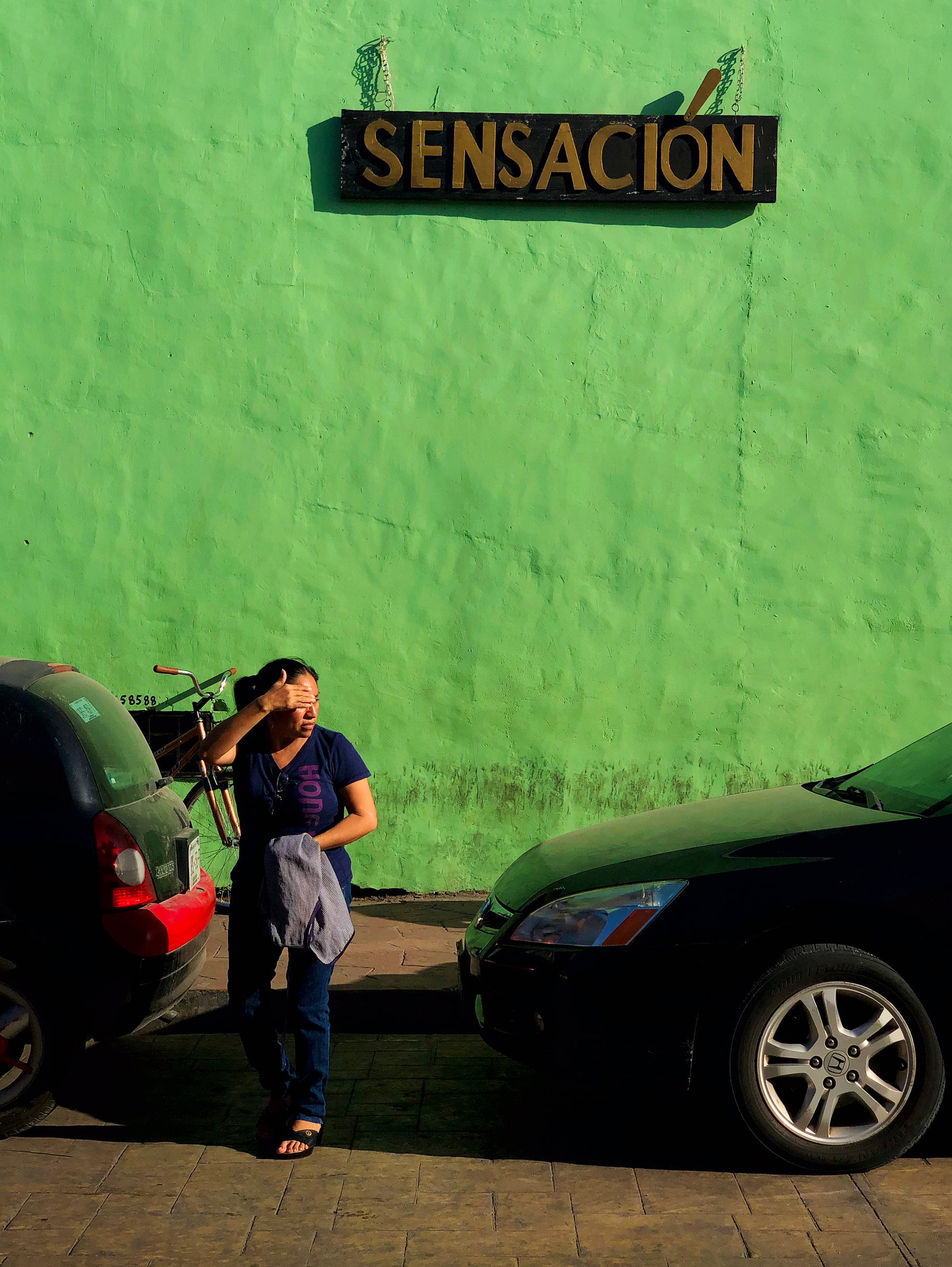 A woman looks out for incoming traffic before crossing the road. Behind her a green wall with a sign that reads “Sensacion”