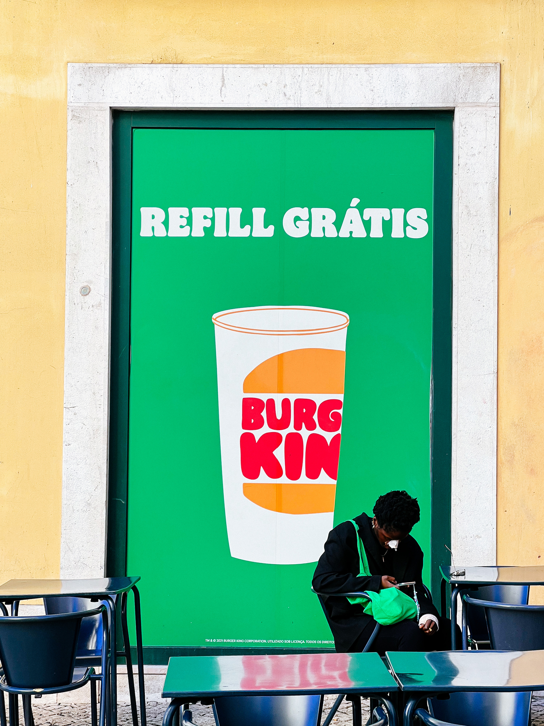 A woman uses her phone in front of a Burger King window, the words “Free Refill” painted on it. 