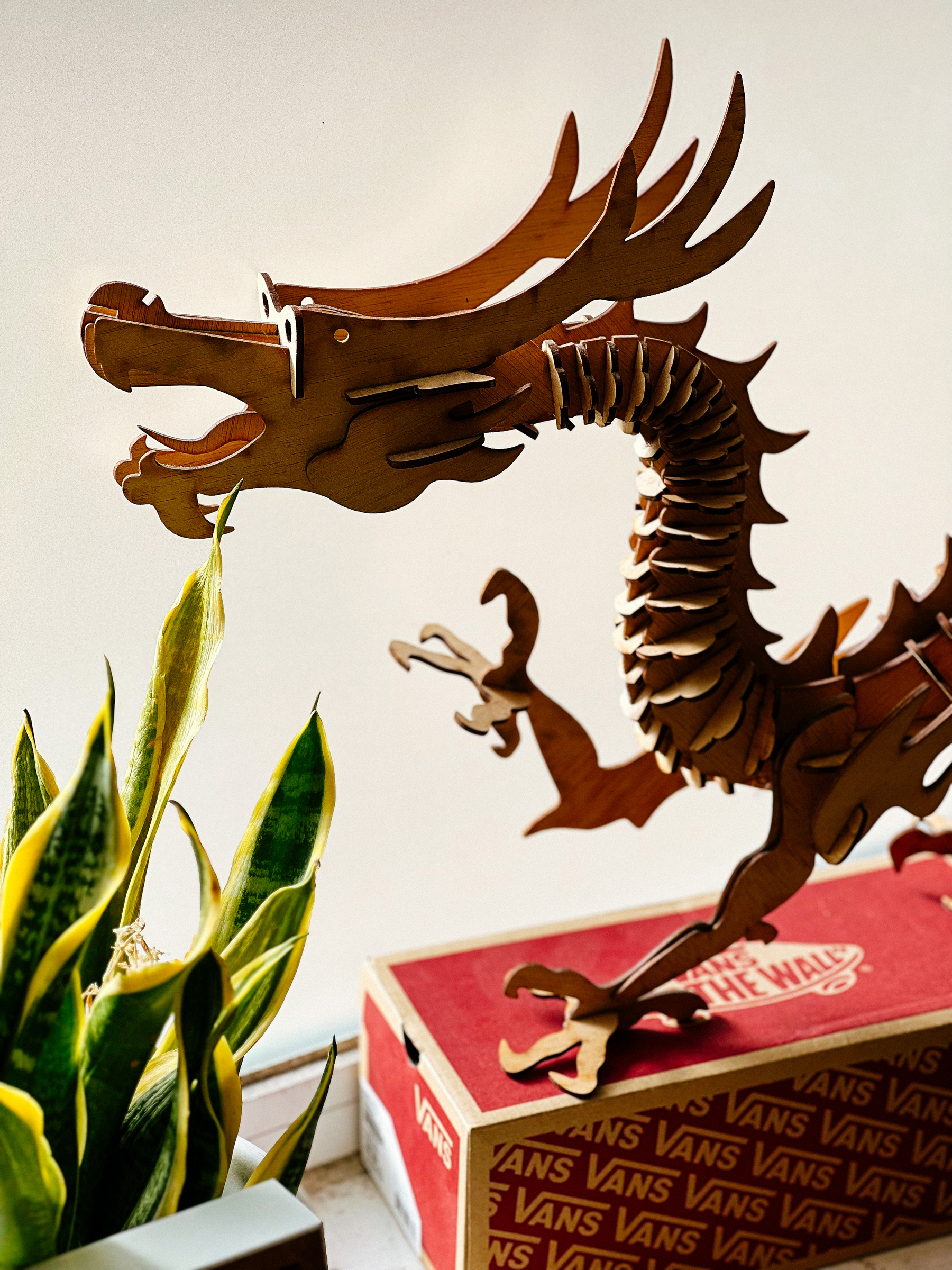 A wooden dragon sits on top of a shoebox, a plant by their side. 