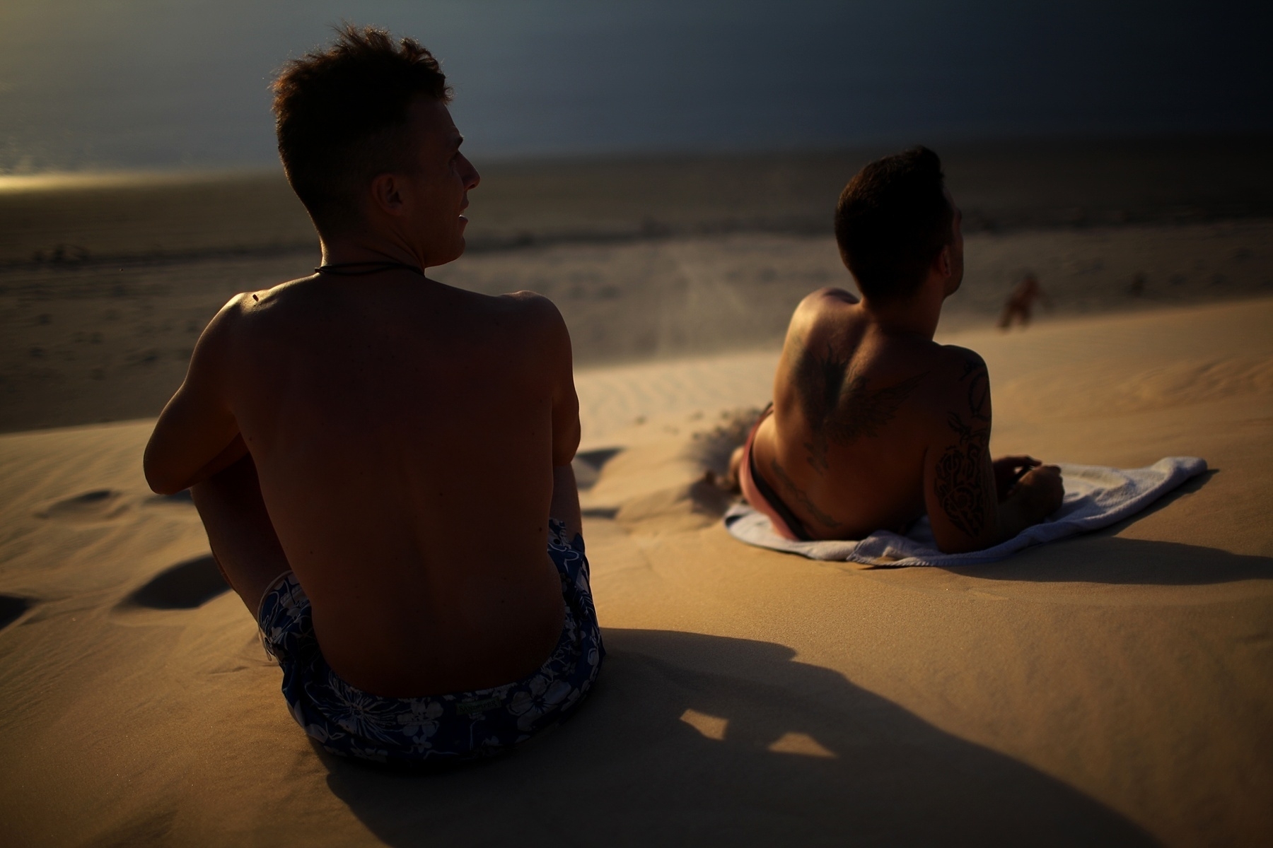 Two man sit on top of a sand dune, watching the sea below.
