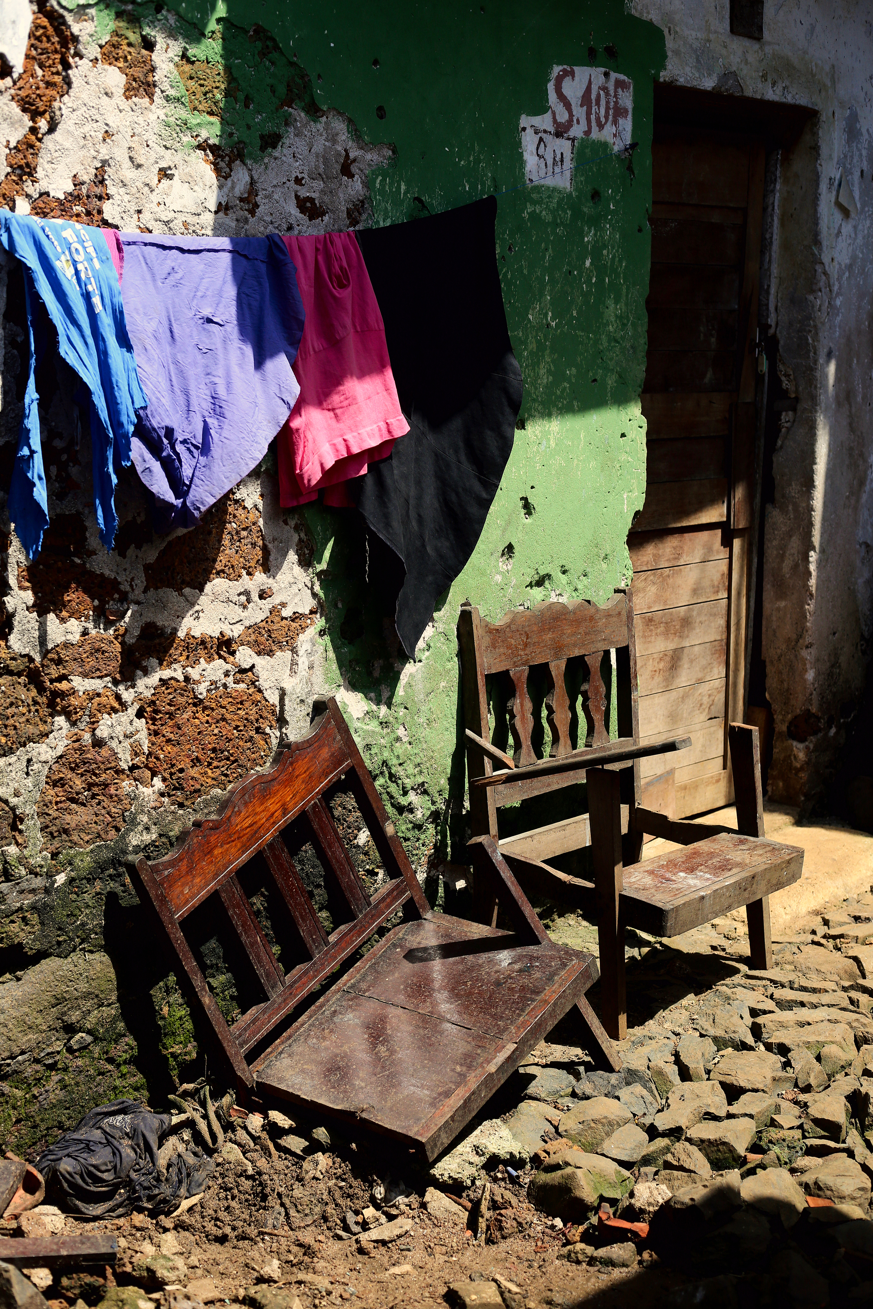 Two completely broken chairs stand outside a house, with clothes hanging from a line above them. 