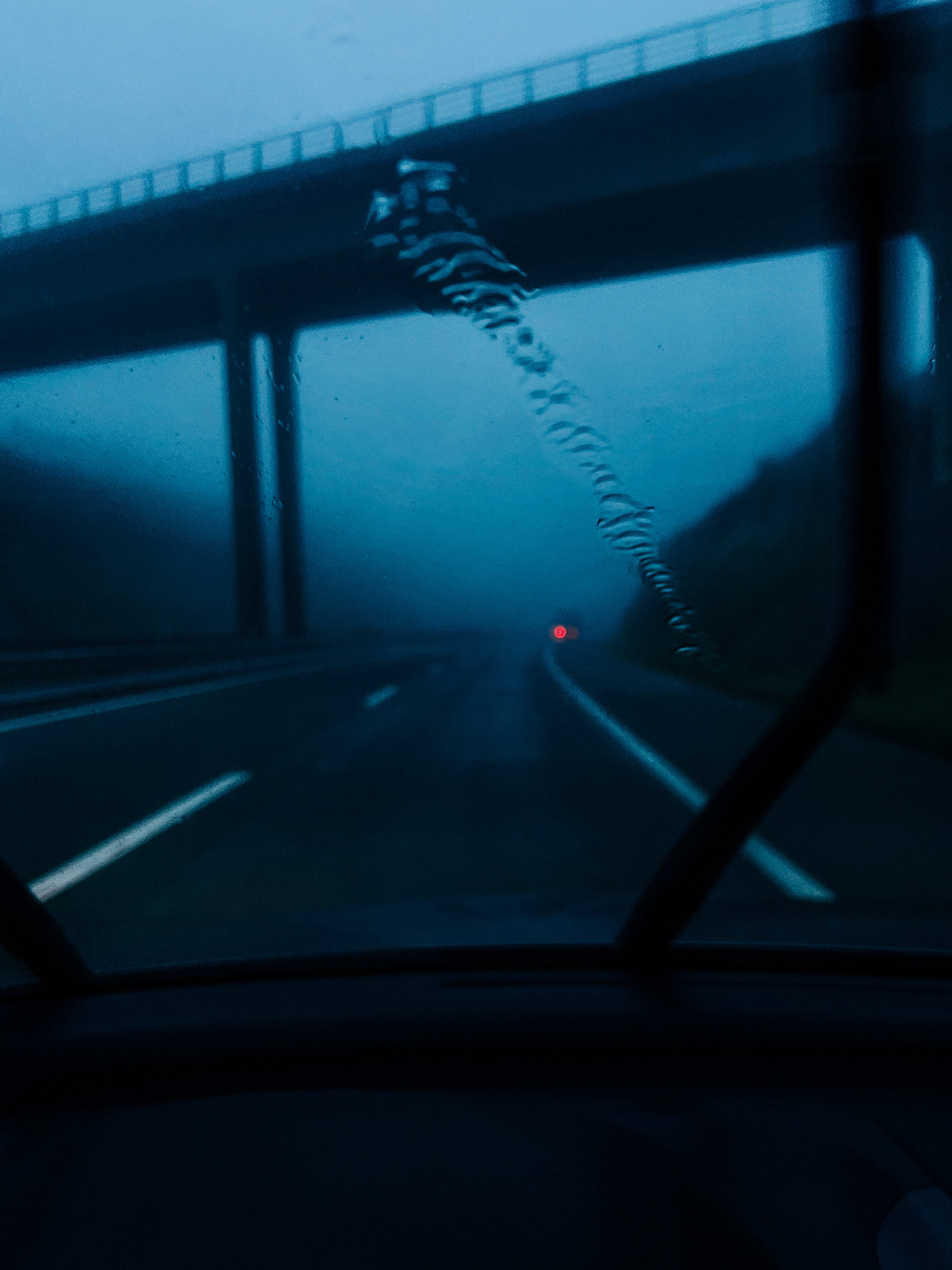 Driving on a dark road, a bridge is seen ahead, and the red light from a car in the distance. 