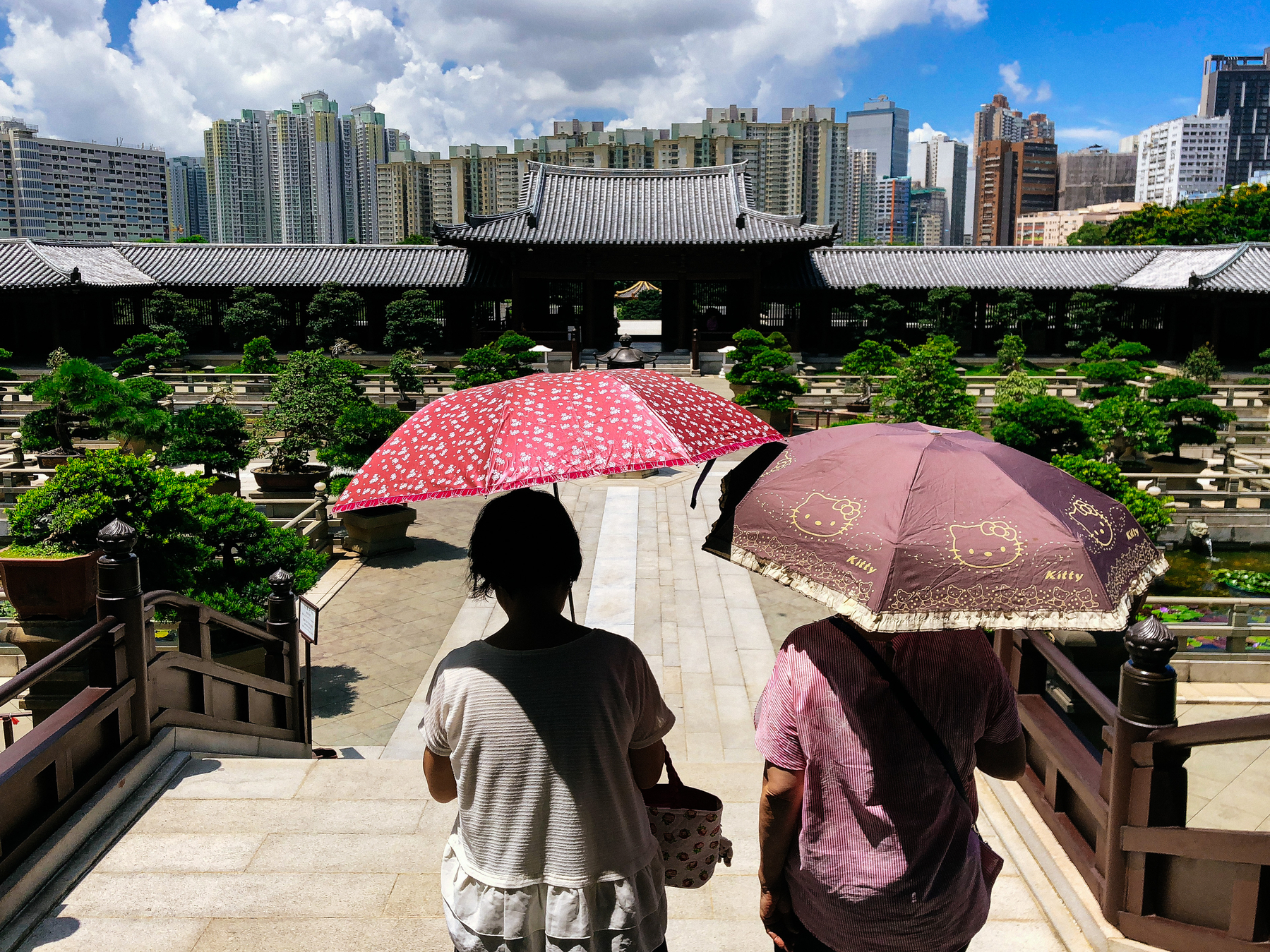 Two women look away from us, looking at a metropolis skyline. Both have umbrellas, one of these has Hello Kitty on it. 