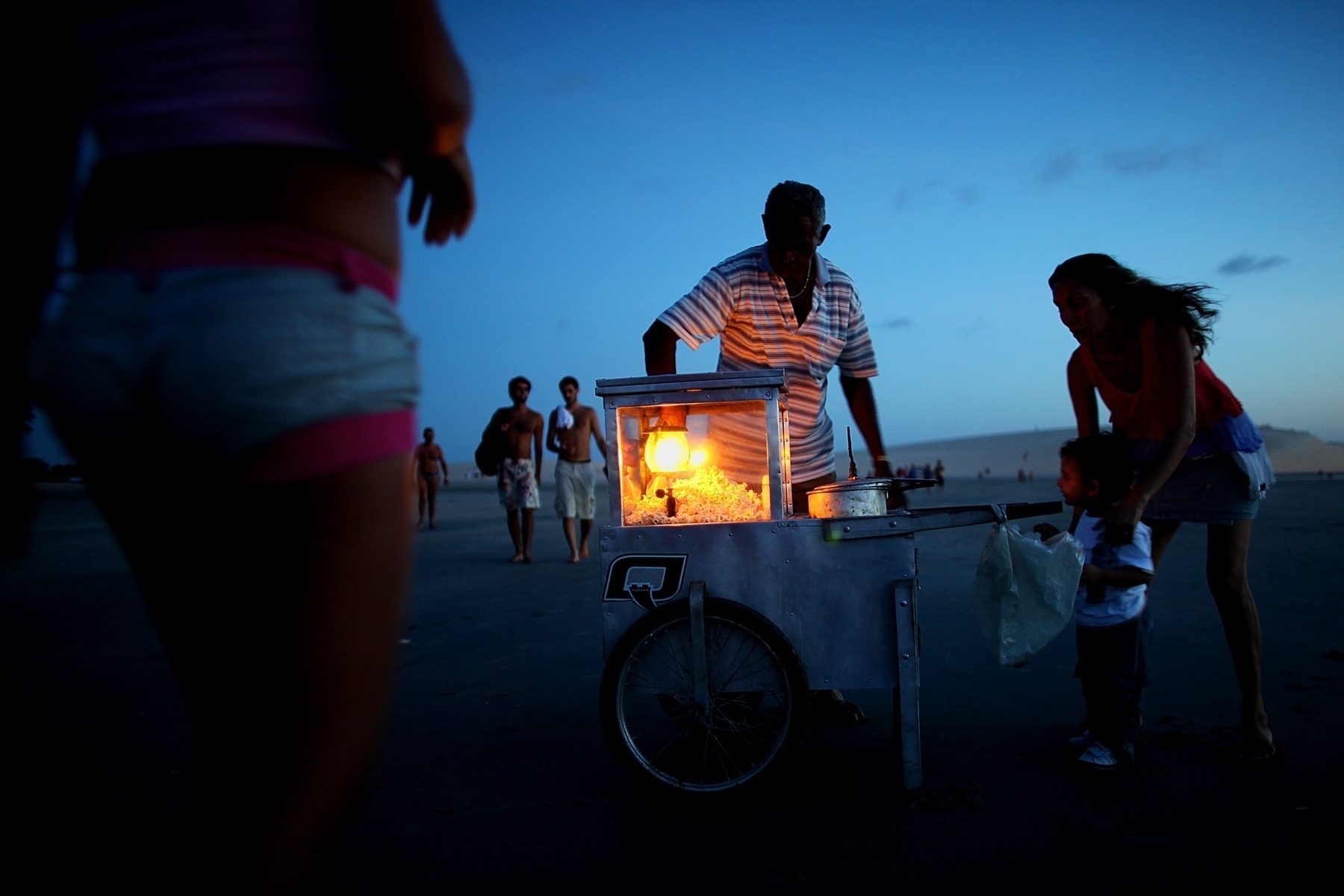 A man sells popcorn at the beach during sunset. As people walk by a mom waits with her boy for the popcorn the man is preparing.
