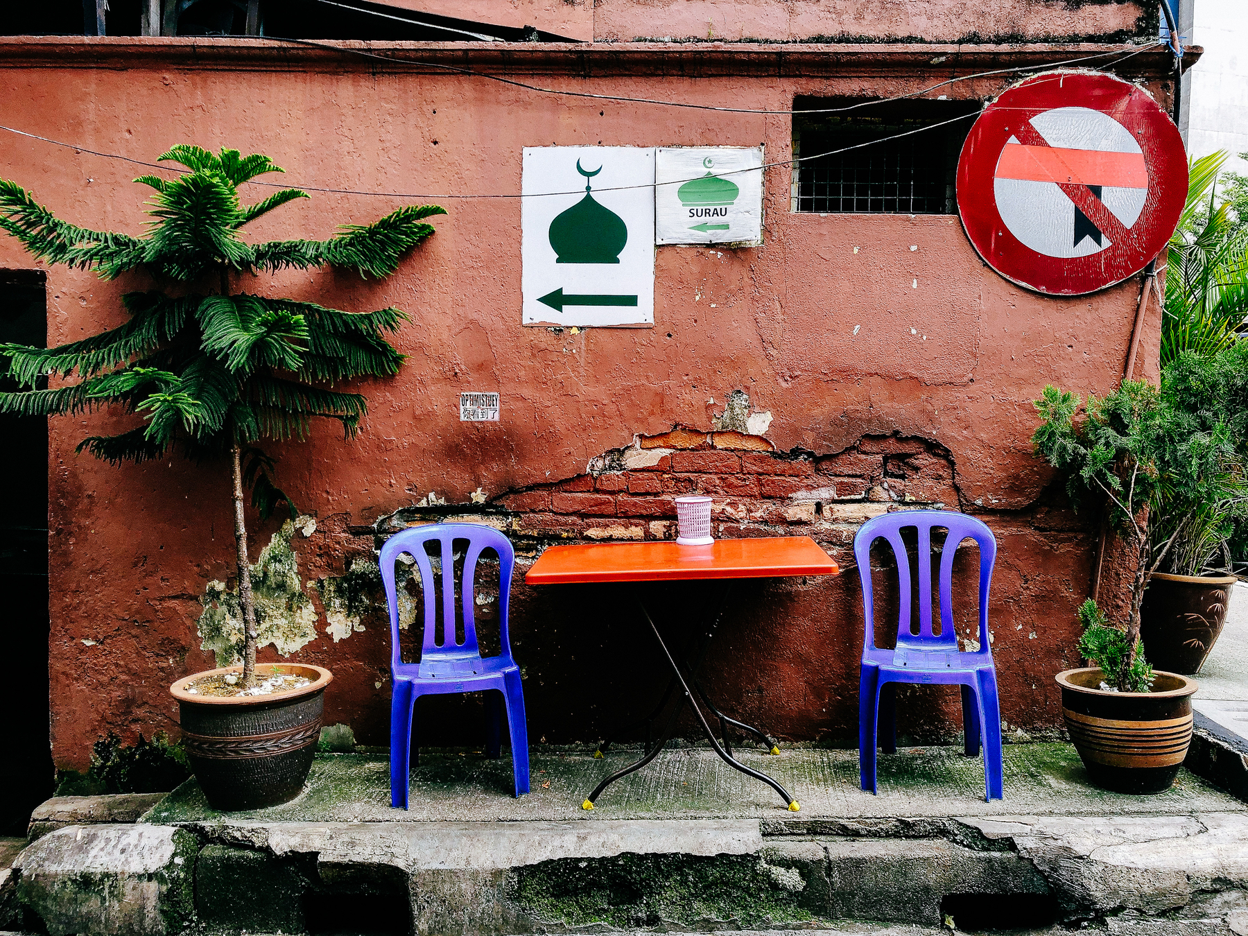 Two plastic chairs and a table, next to a wall where signs directing people to a mosque are posted. 